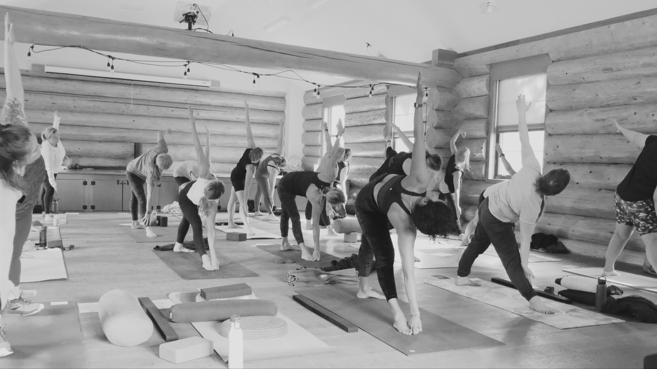 People participating in a yoga class in a log cabin studio, practicing side stretches on yoga mats with props, under string lights, with windows on wooden walls.