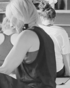 Close-up of a woman with blonde hair in a bun, wearing a sleeveless top, seated next to another person with dark hair, in an indoor setting.