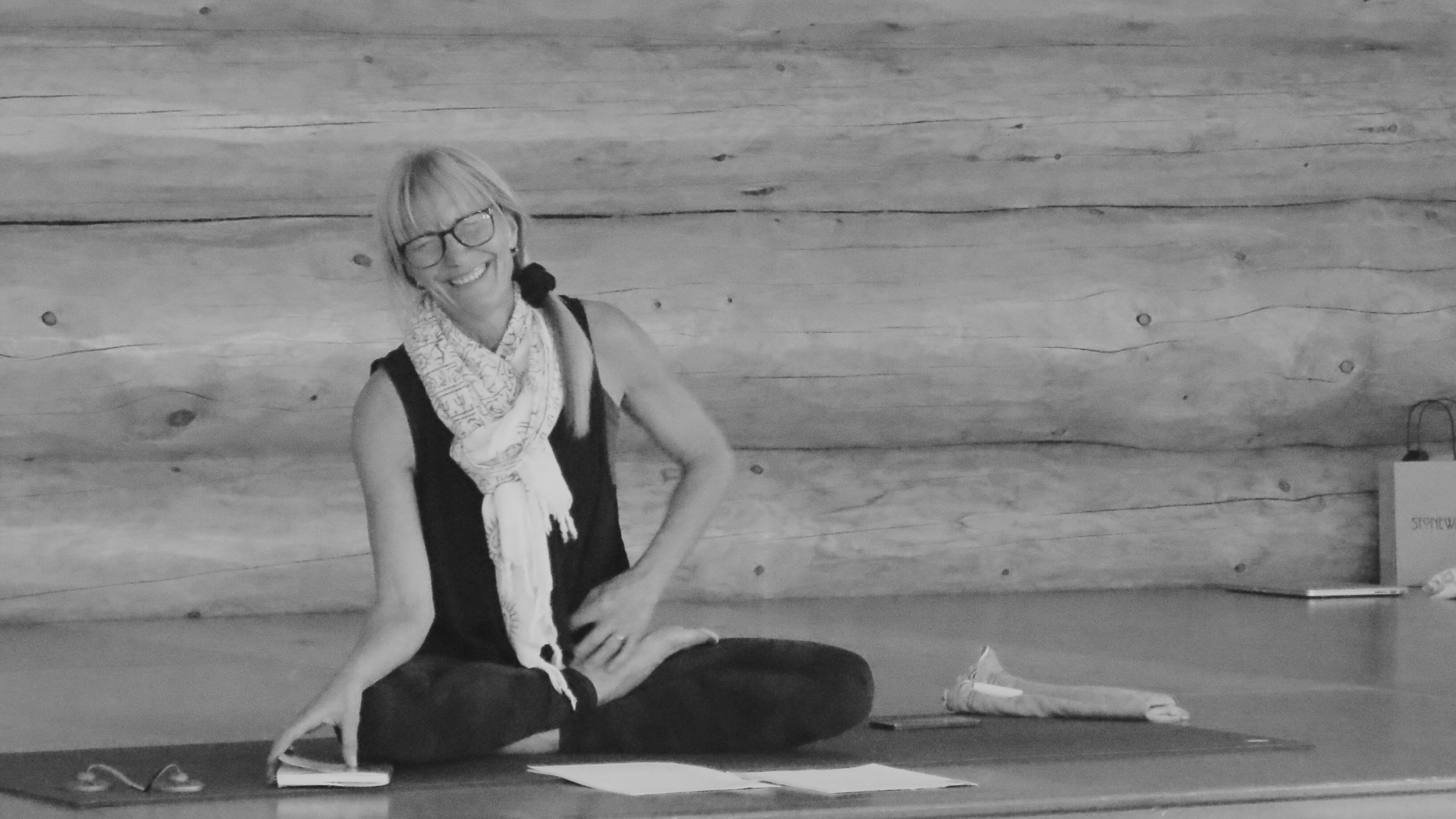 A woman with glasses and blonde hair, sitting cross-legged on the floor, smiling, with papers and a notebook in front of her, against a wooden wall.