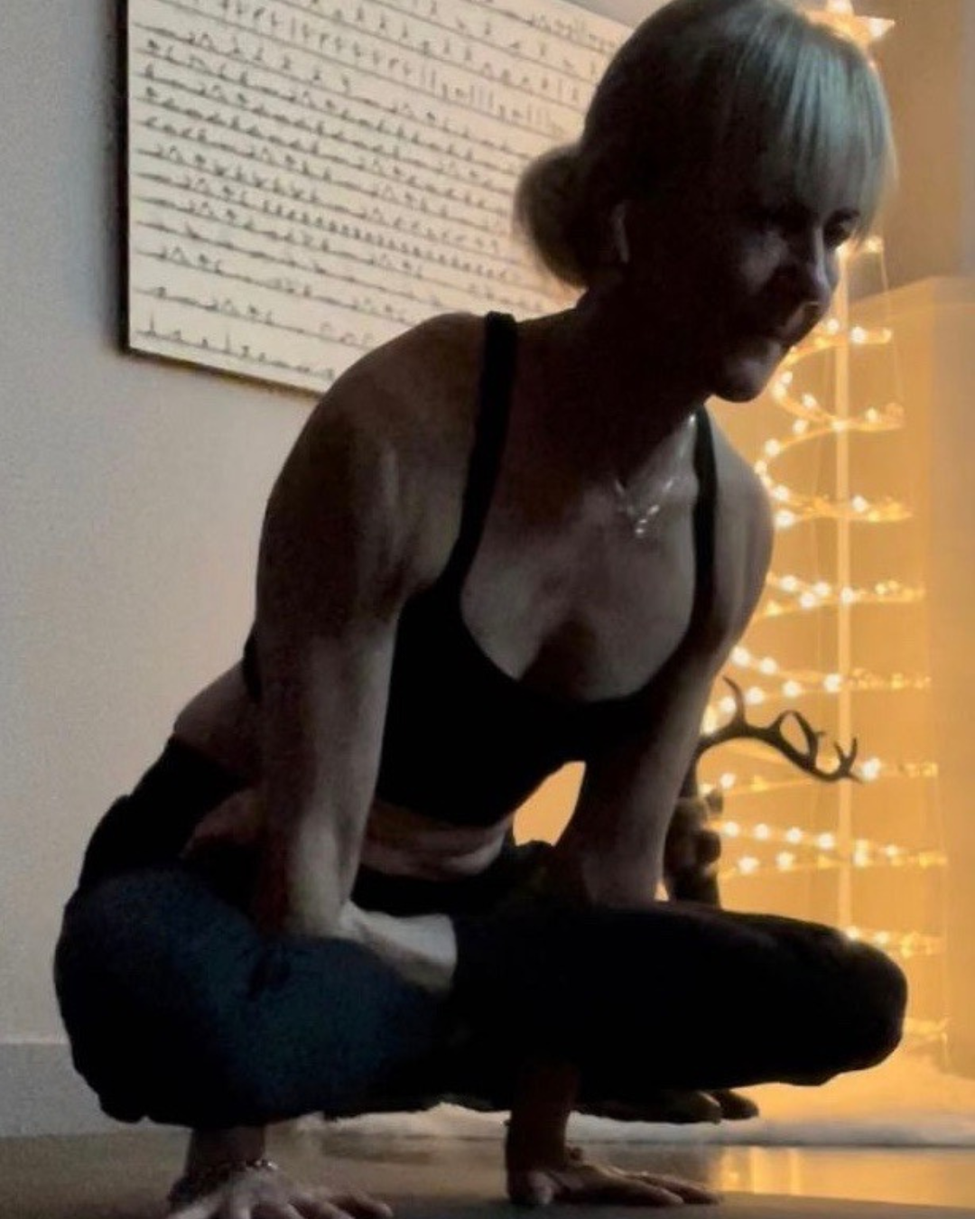 A woman practicing yoga indoors, in a crouched position with hands on the floor. Behind her, a painting with horizontal lines and a wooden reindeer decor with lights are visible.