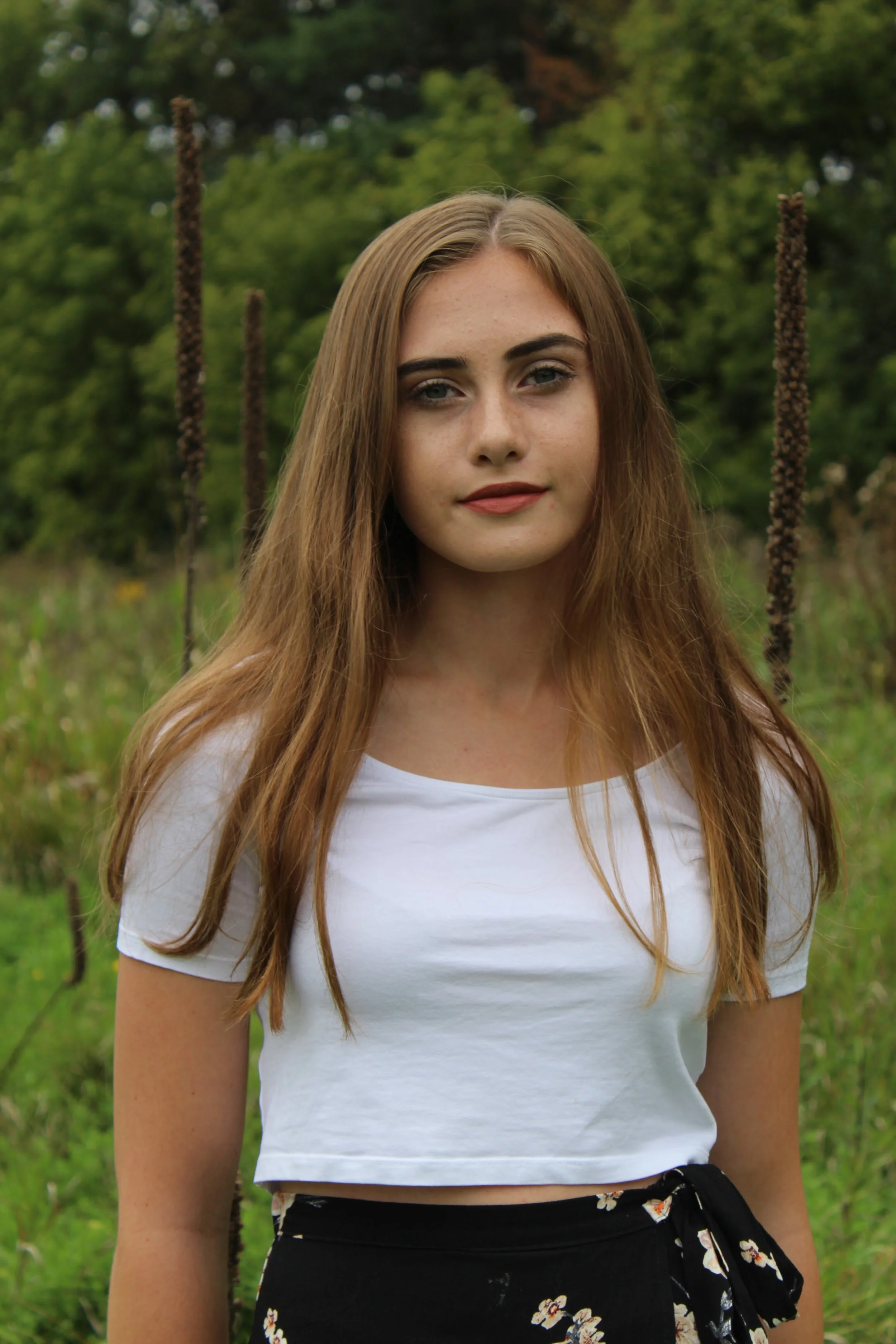 A young woman with long, light brown hair and fair skin, standing outdoors in a grassy field with greenery in the background. She is wearing a white cropped t-shirt and a black skirt with a floral pattern.