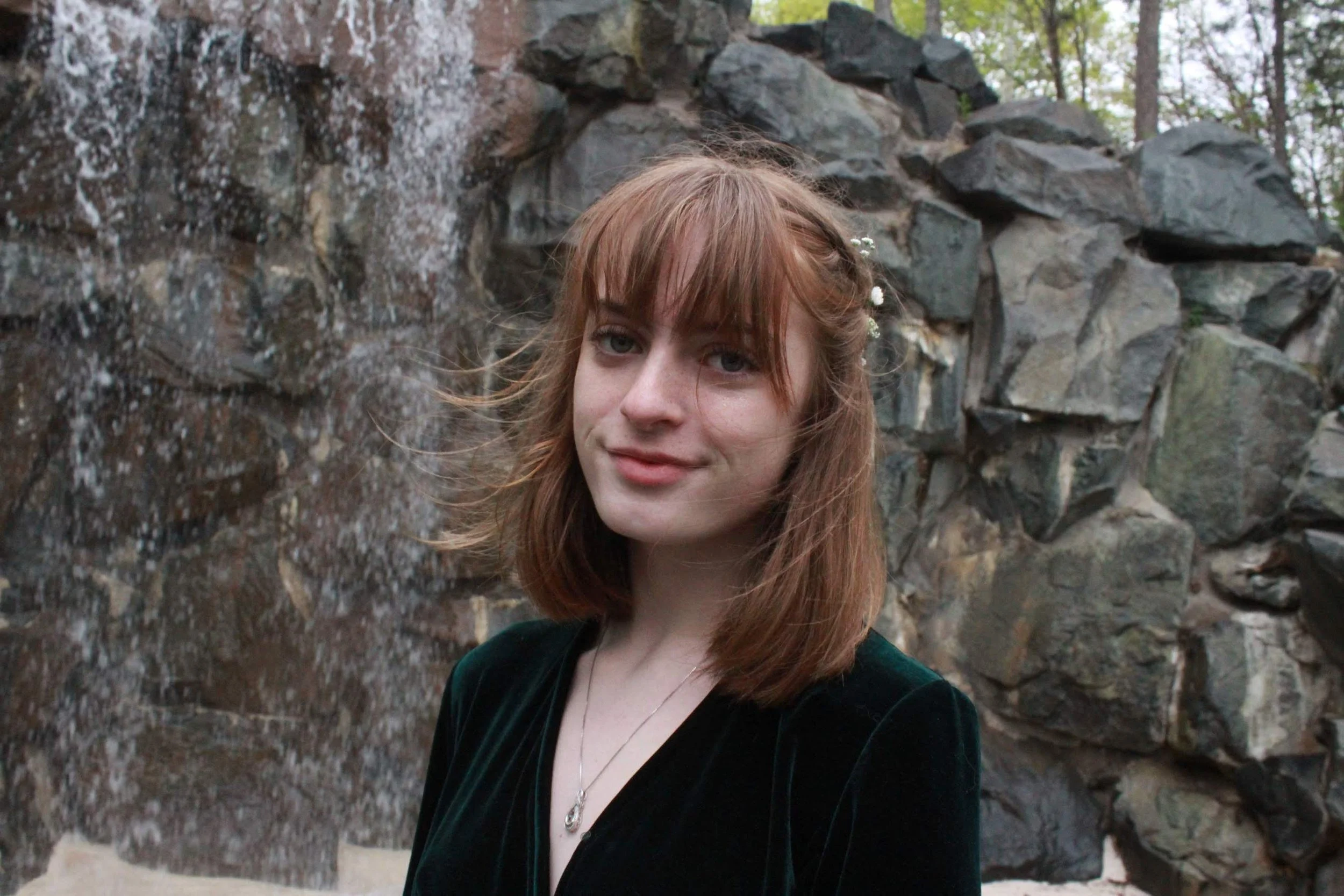 A young woman with shoulder-length red hair, wearing a black top and a necklace, standing outdoors in front of a rocky waterfall and trees.