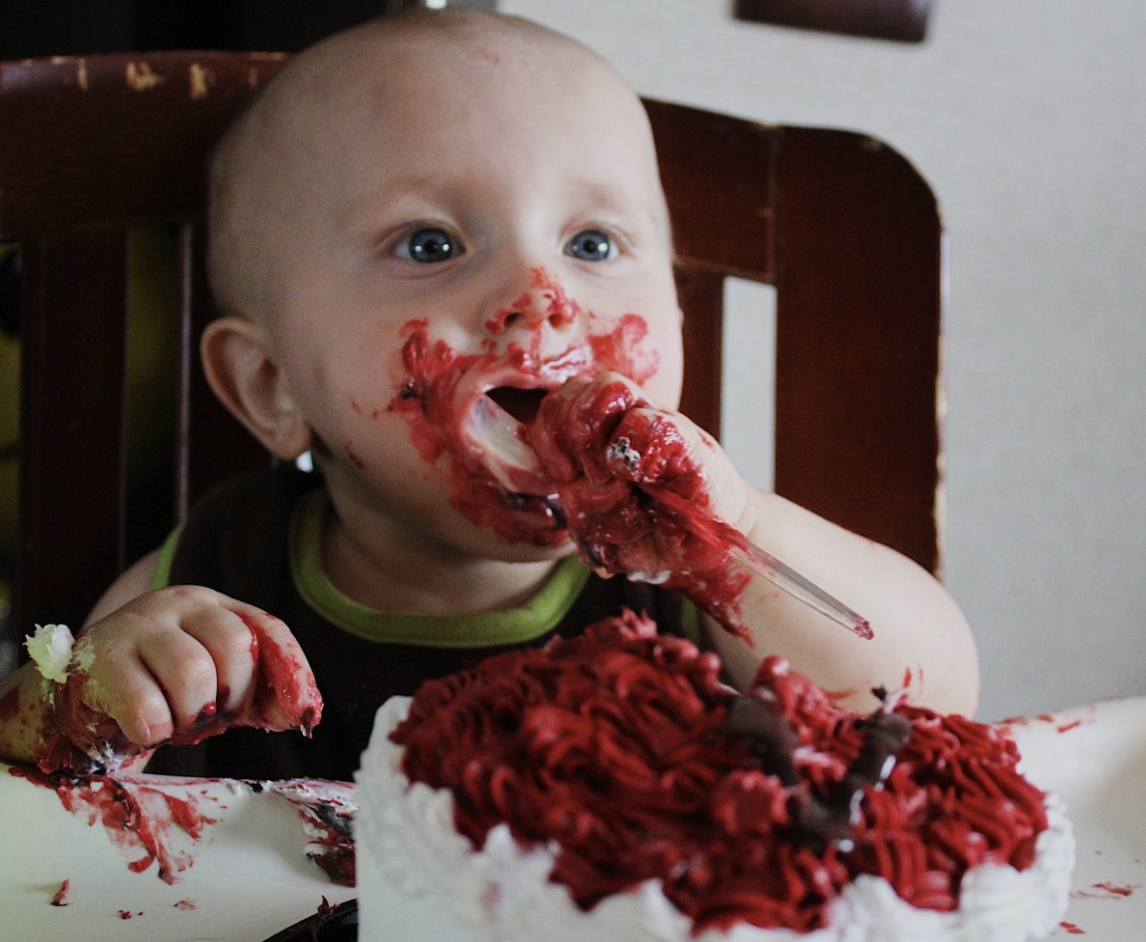 A young child with a messy face and hands covered in red frosting, eating from a birthday cake with red and white frosting and chocolate decorations.