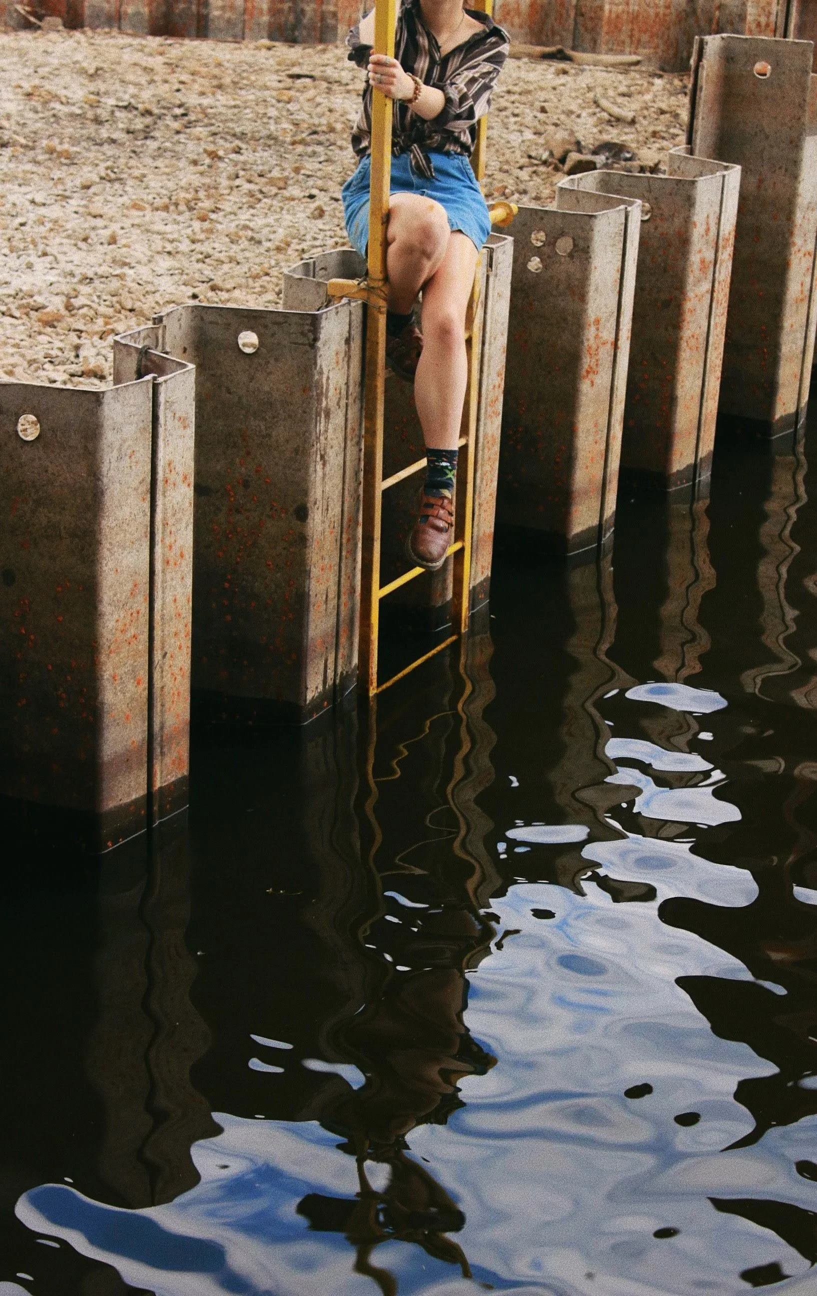 Person sitting on a yellow platform with legs dangling over water, hanging from a metal ladder, near a rusted metal wall and muddy ground.