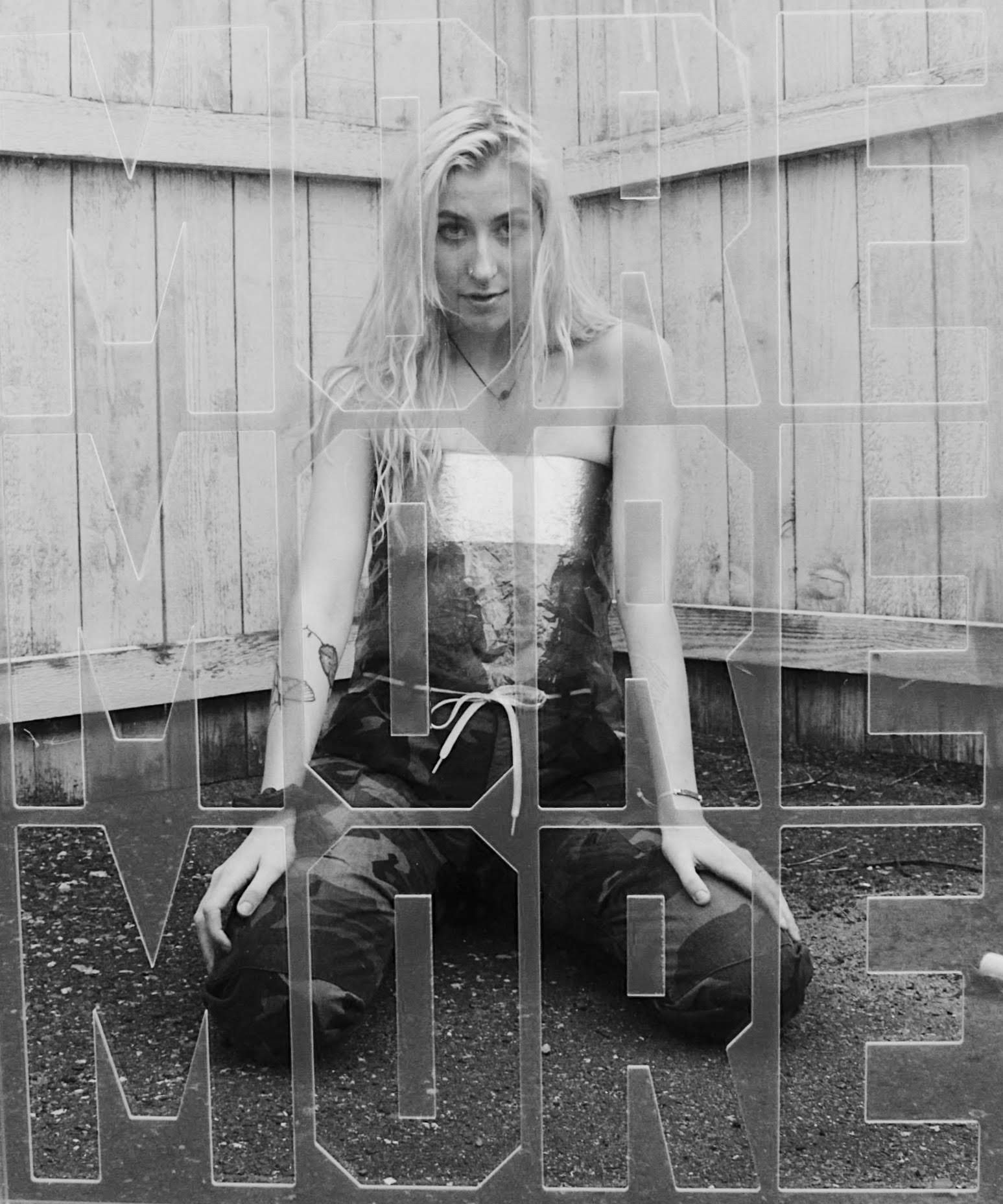 A young woman with long blonde hair sitting on the ground in front of a wooden fence, wearing a strapless top, camouflage pants, and a necklace, looking at the camera.