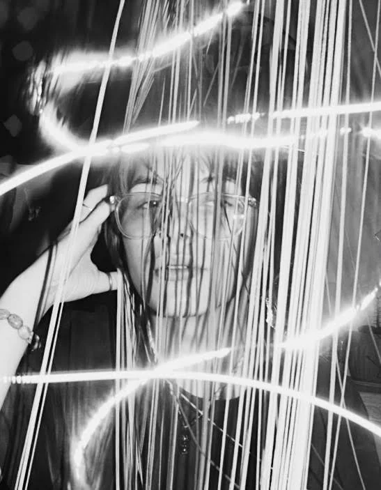 Black and white photo of a woman smiling behind light streaks and hanging strings or lights, wearing glasses.
