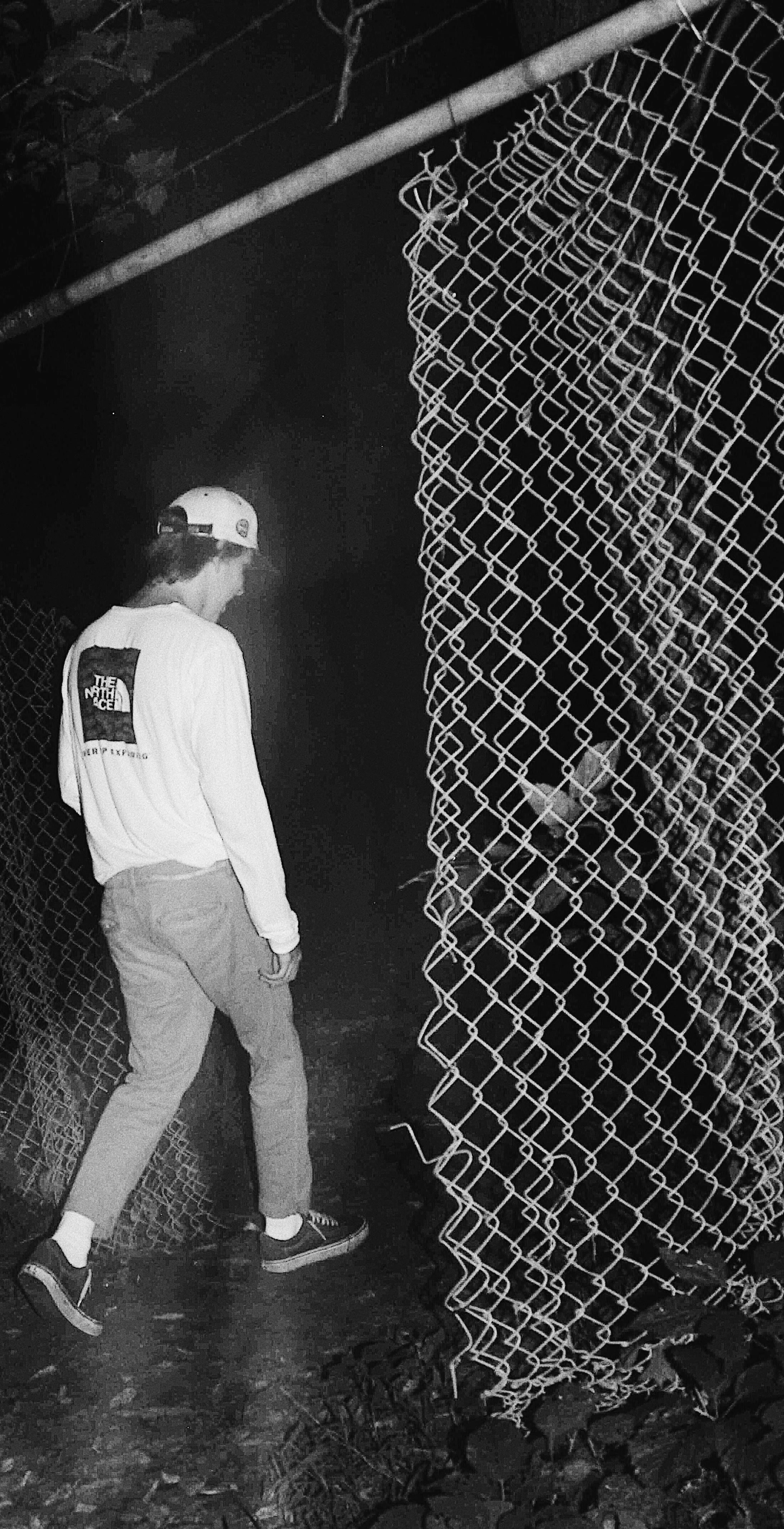 A person in casual clothing, including a cap with a logo, stands next to a chain-link fence at night. The person appears to be looking at or near a large, tangled roll of wire mesh leaning against the fence.