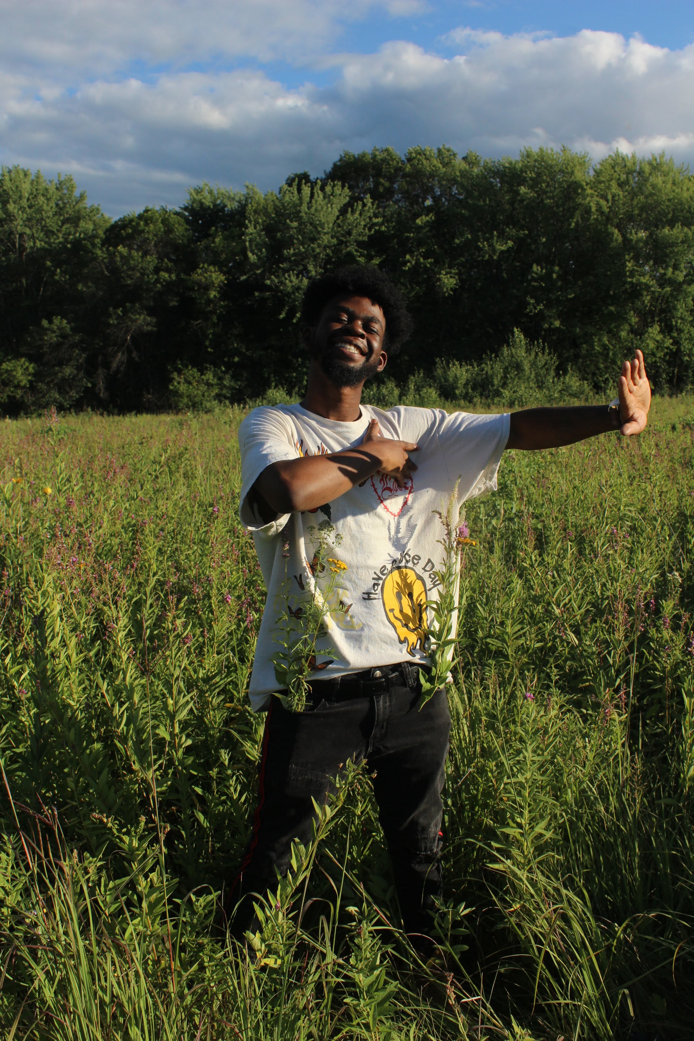 A smiling man stands in a green field with tall grass and wildflowers, pointing to his chest with one hand and raising the other arm with an open palm, under a cloudy sky with sun rays.