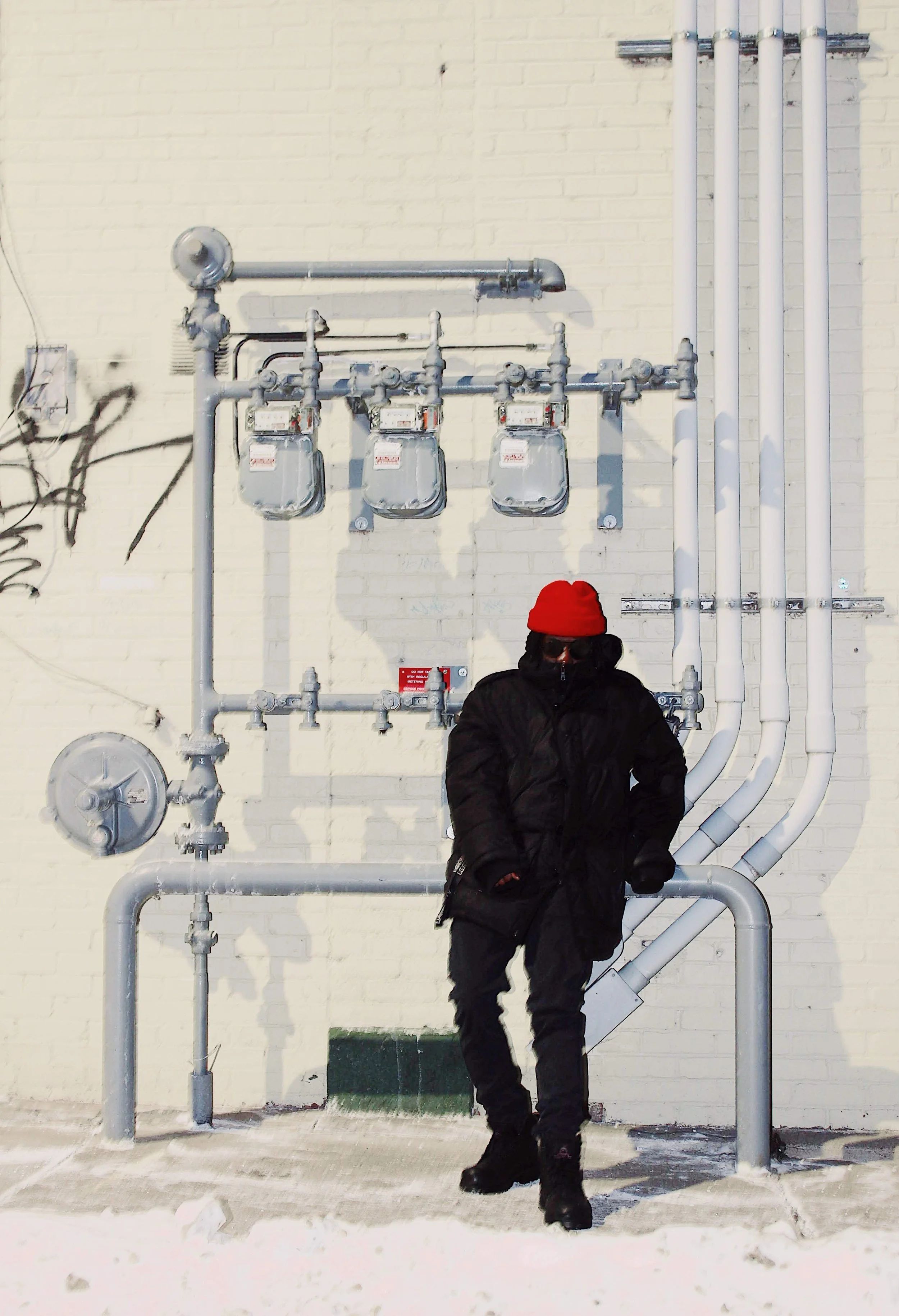 Person dressed in black winter coat, red beanie, and sunglasses standing in front of industrial pipes and meters on a white brick wall, with snow on the ground.
