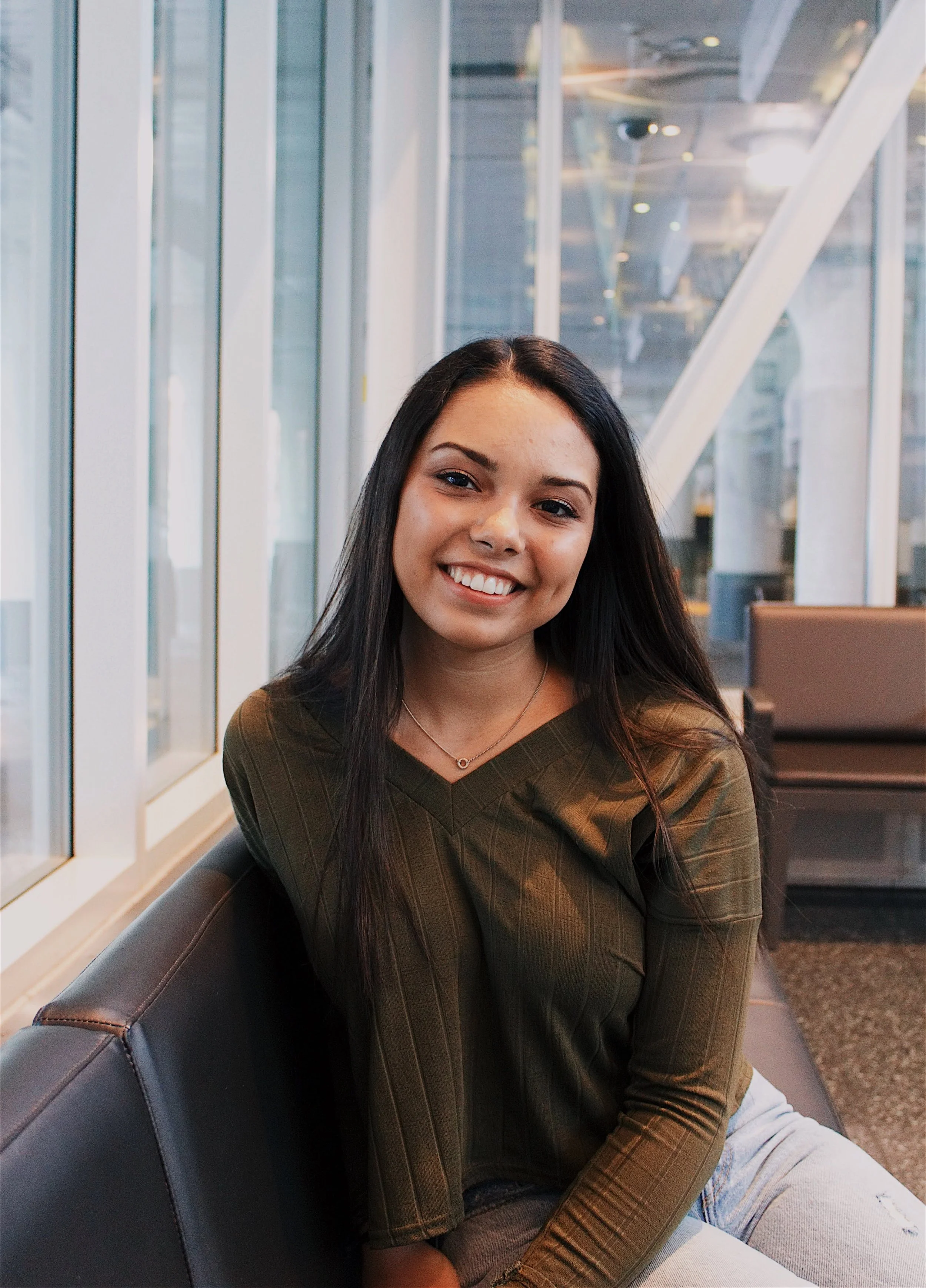 A young woman with long dark hair smiling, sitting on a bench in a modern indoor space with large glass windows and metal structures.