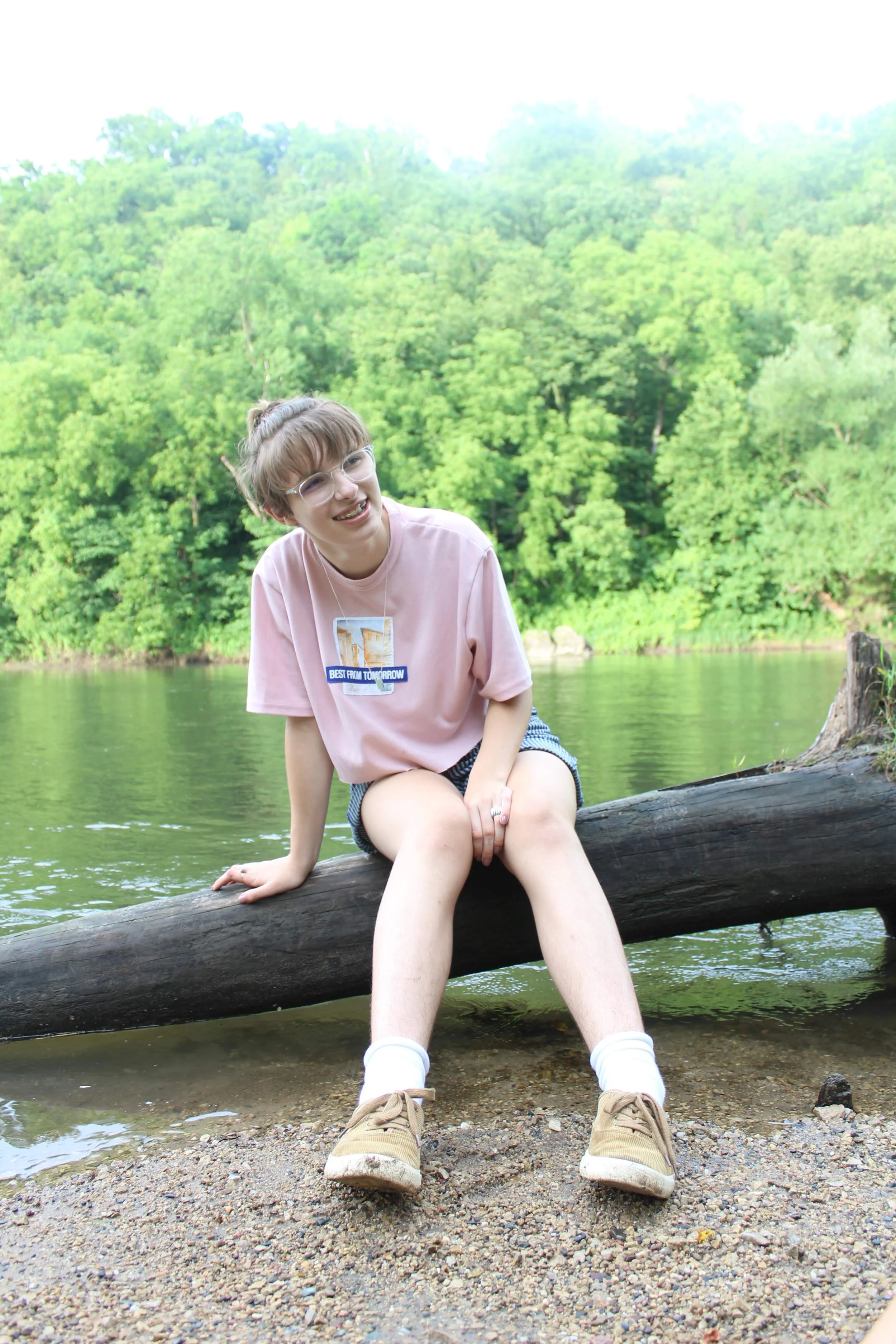 A young woman sitting on a fallen log by a river, smiling, with green trees in the background.