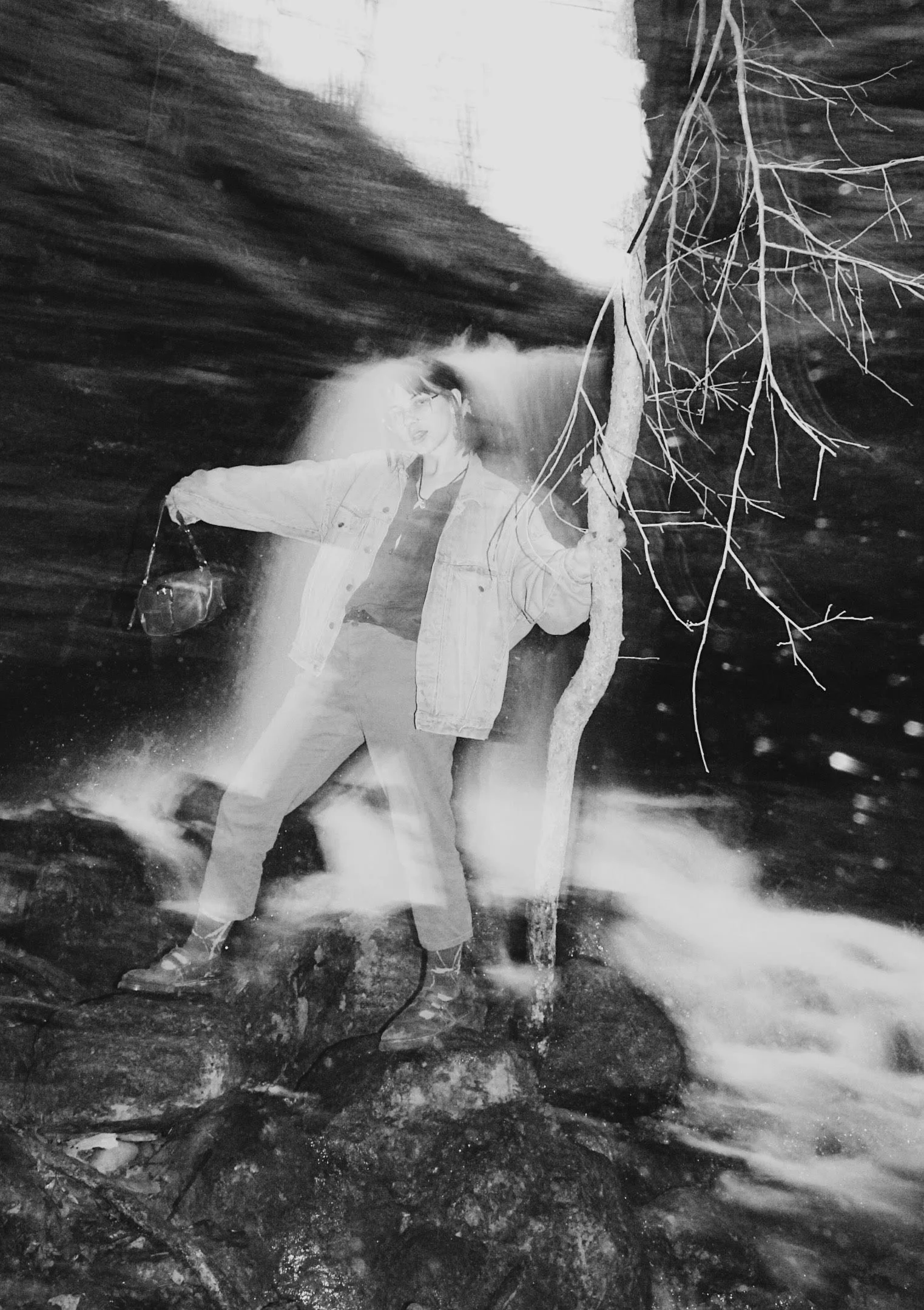A person standing on rocks near a creek or river, holding a camera in one hand, with a tree next to them. The photo is in black and white, with a long exposure effect showing motion blur.