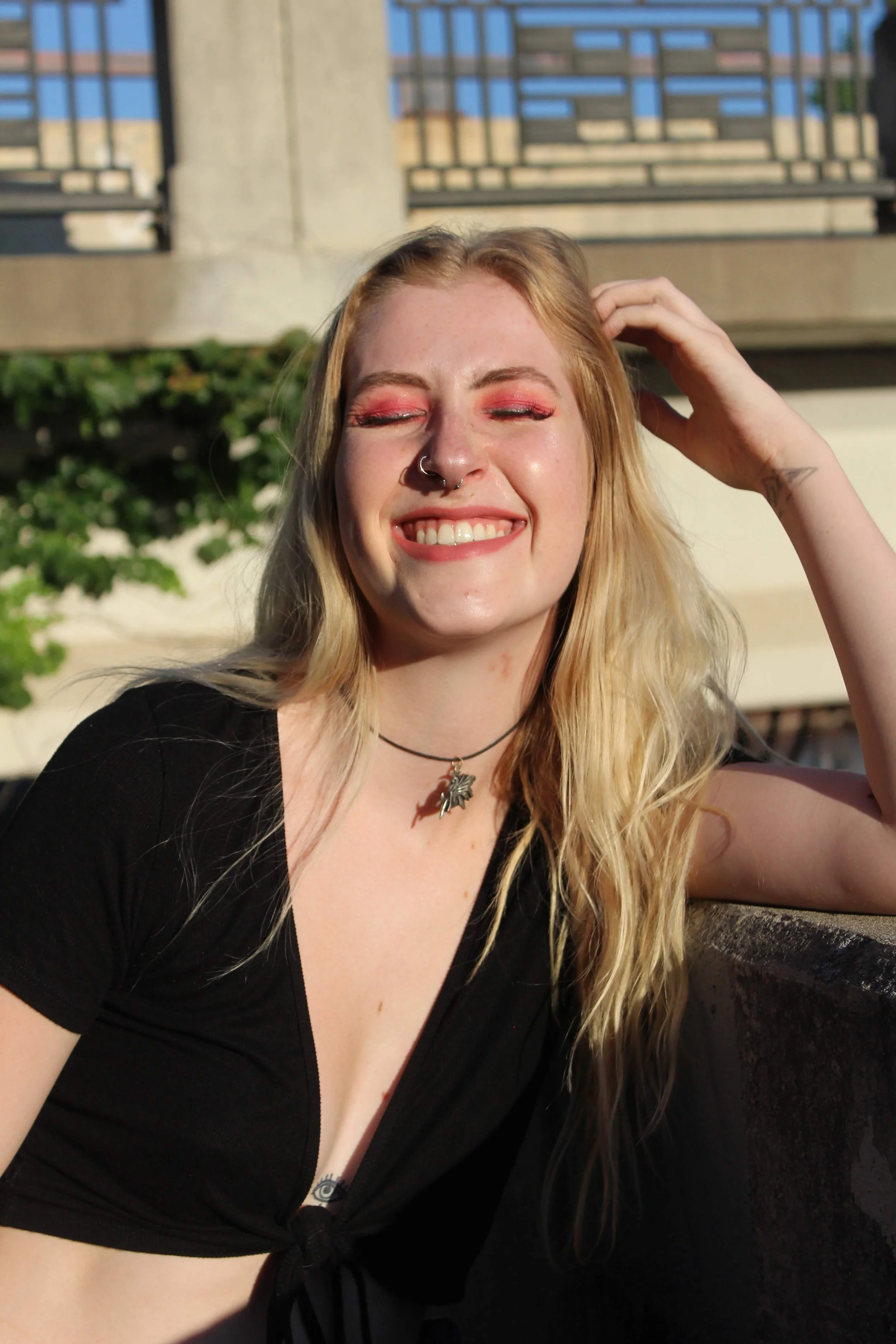 A young woman with long blonde hair, wearing a black top, smiling with eyes closed, and posing outdoors in sunlight near a railing and greenery.
