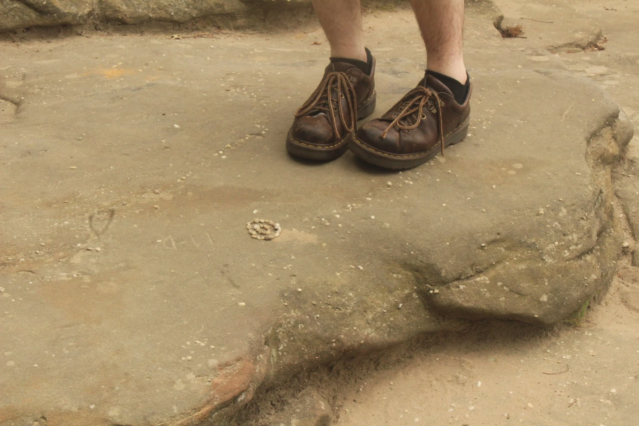 Close-up of a person’s feet in worn brown leather shoes standing on a large flat rock with engraved initials and symbols on dirt ground.