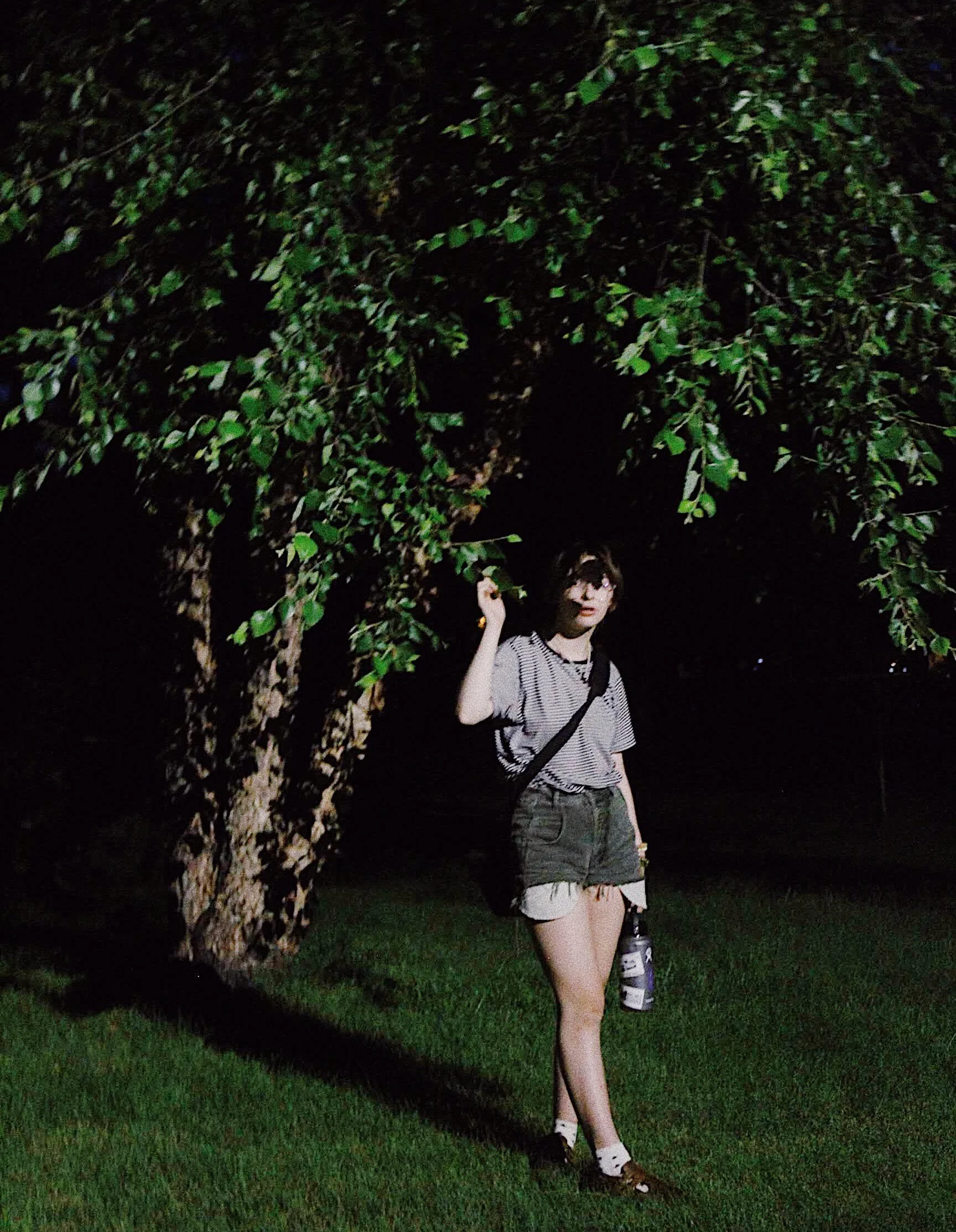 A young woman with dark hair, wearing glasses, a striped t-shirt, denim shorts, and carrying a backpack, standing under a large leafy tree at night, holding a water bottle.