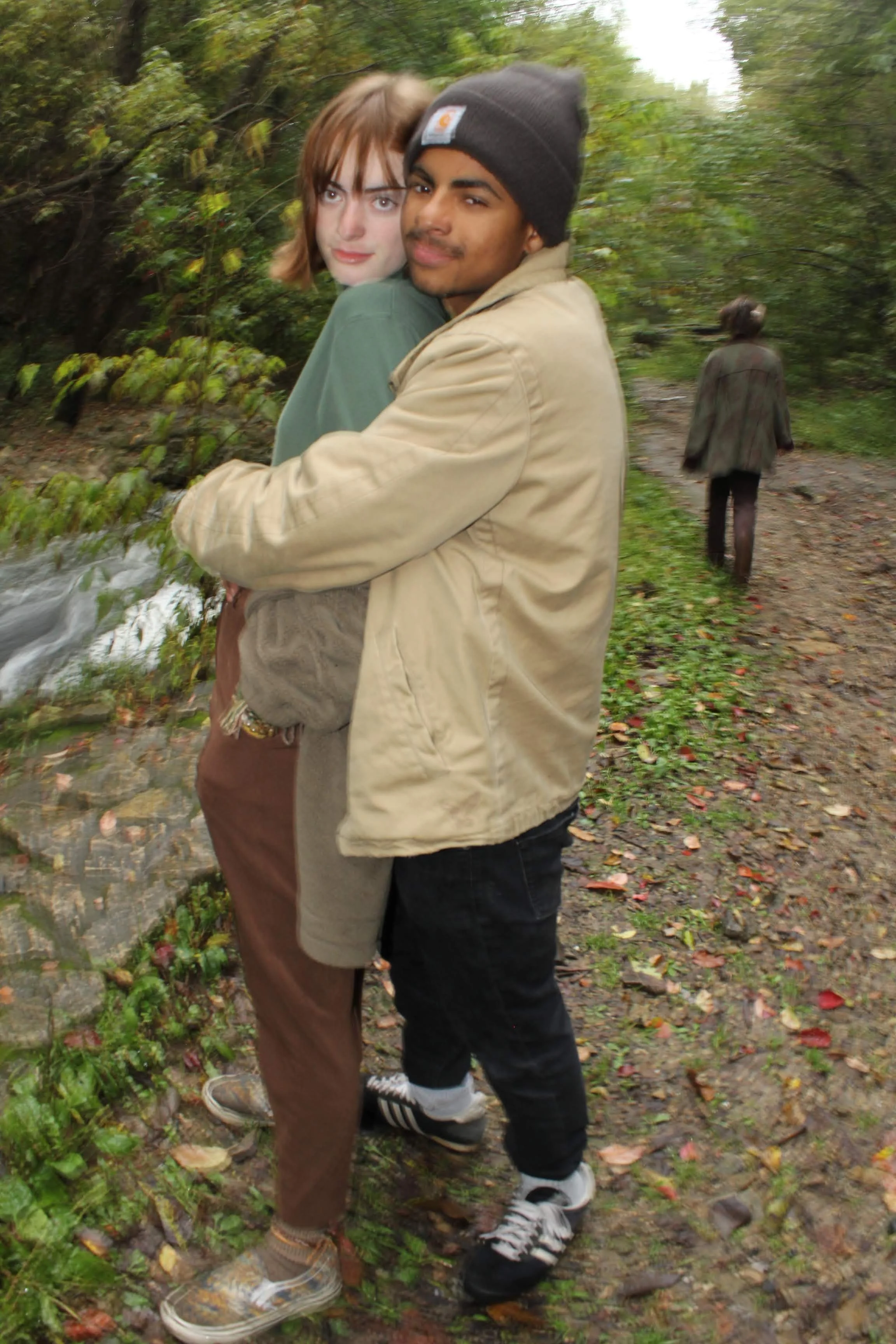 A young couple stands close together on a forest path, with the woman leaning against the man who is wearing a beige jacket and a black beanie. In the background, an older person with gray hair and a brown jacket is walking away along the trail. The 