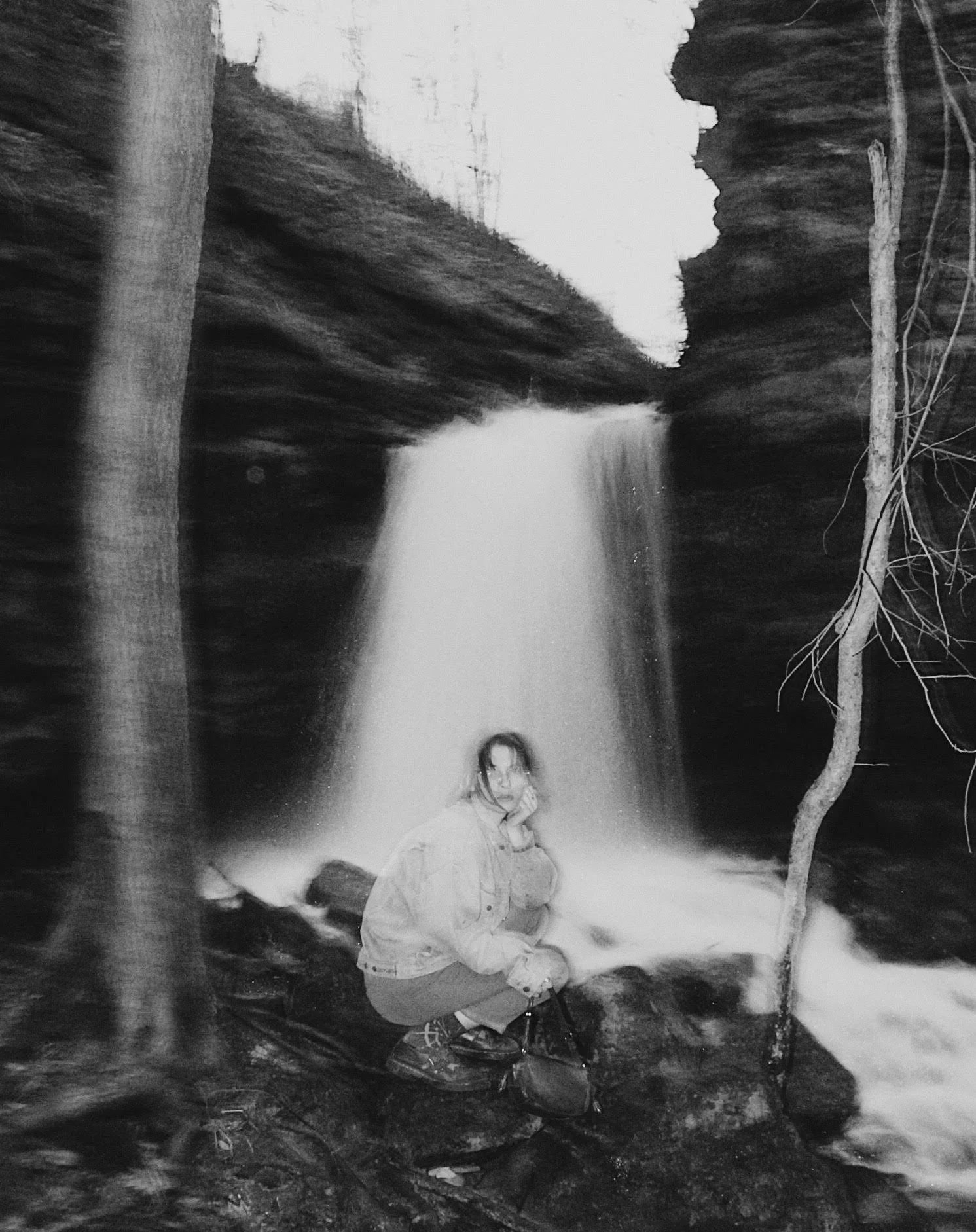 A person sitting on rocks near a waterfall in a natural setting, with trees on either side.