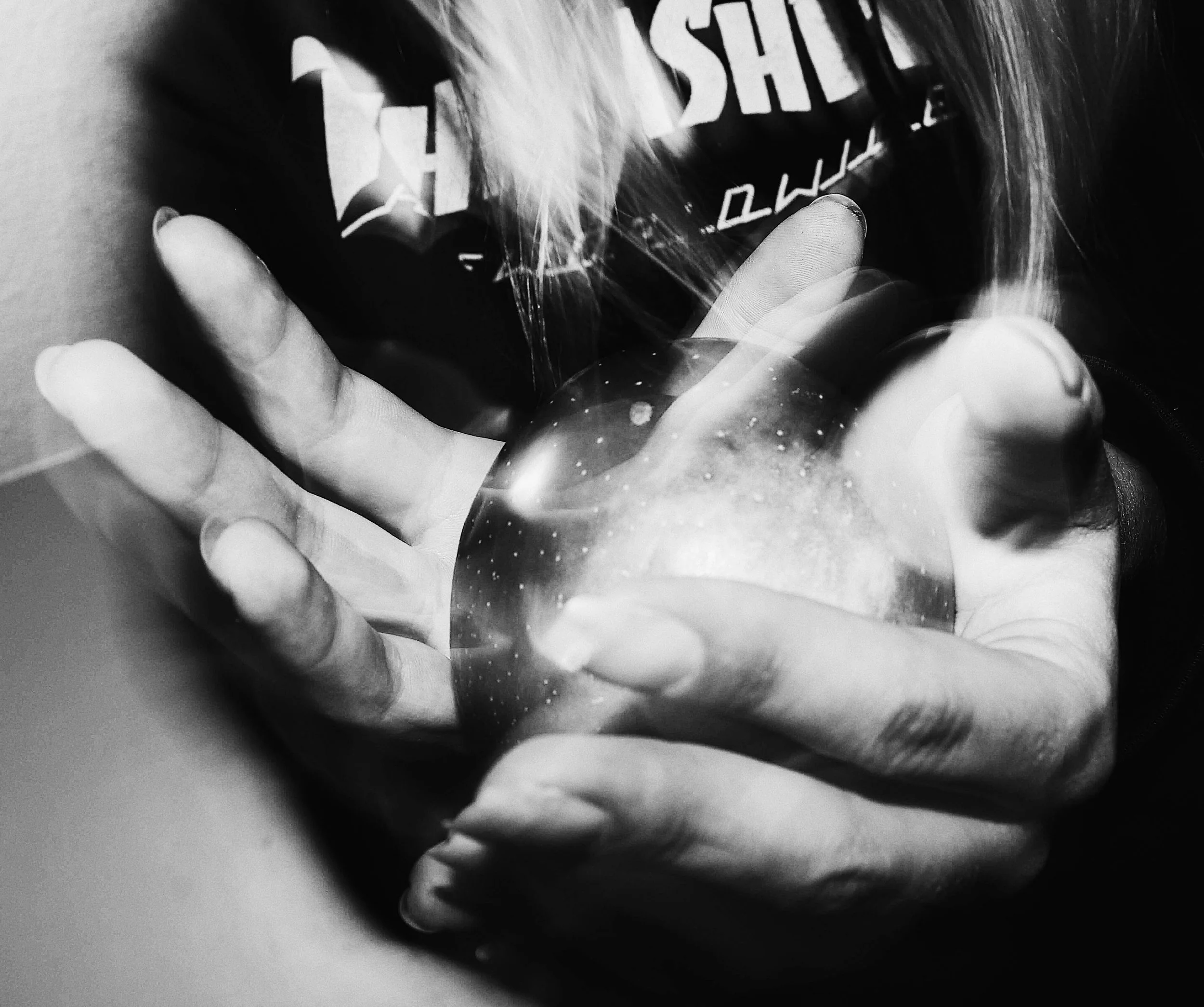 A hand holding a glass orb, with a blurry background featuring a person with long hair and partially visible text on their clothing.