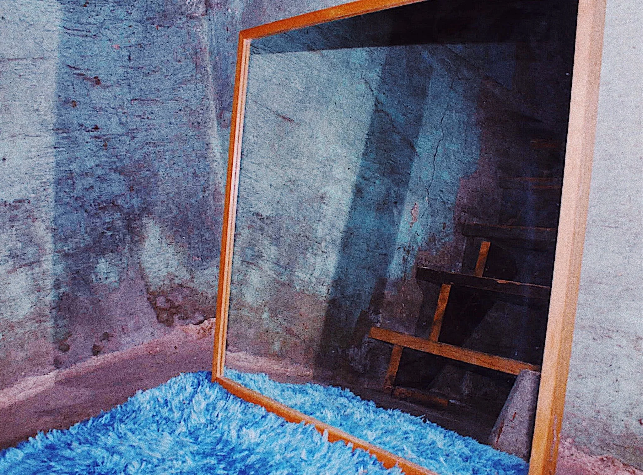 A wooden framed mirror leaning against a rough concrete wall, reflecting a wooden staircase and a blue shaggy rug on the floor.