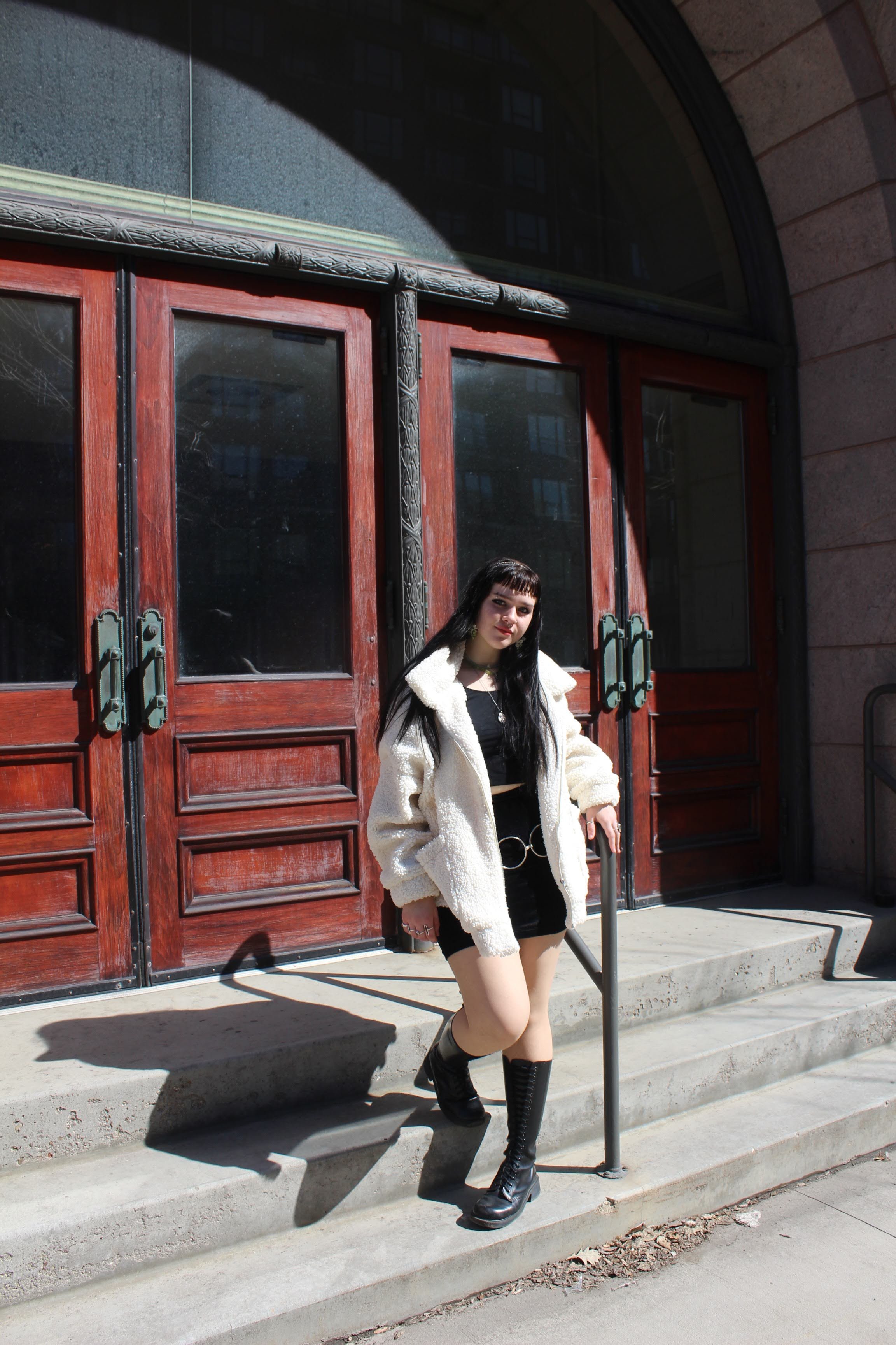 A woman with long dark hair, wearing a white fuzzy jacket, black top and skirt, and black lace-up boots, standing on the steps in front of a wooden door with glass panels.