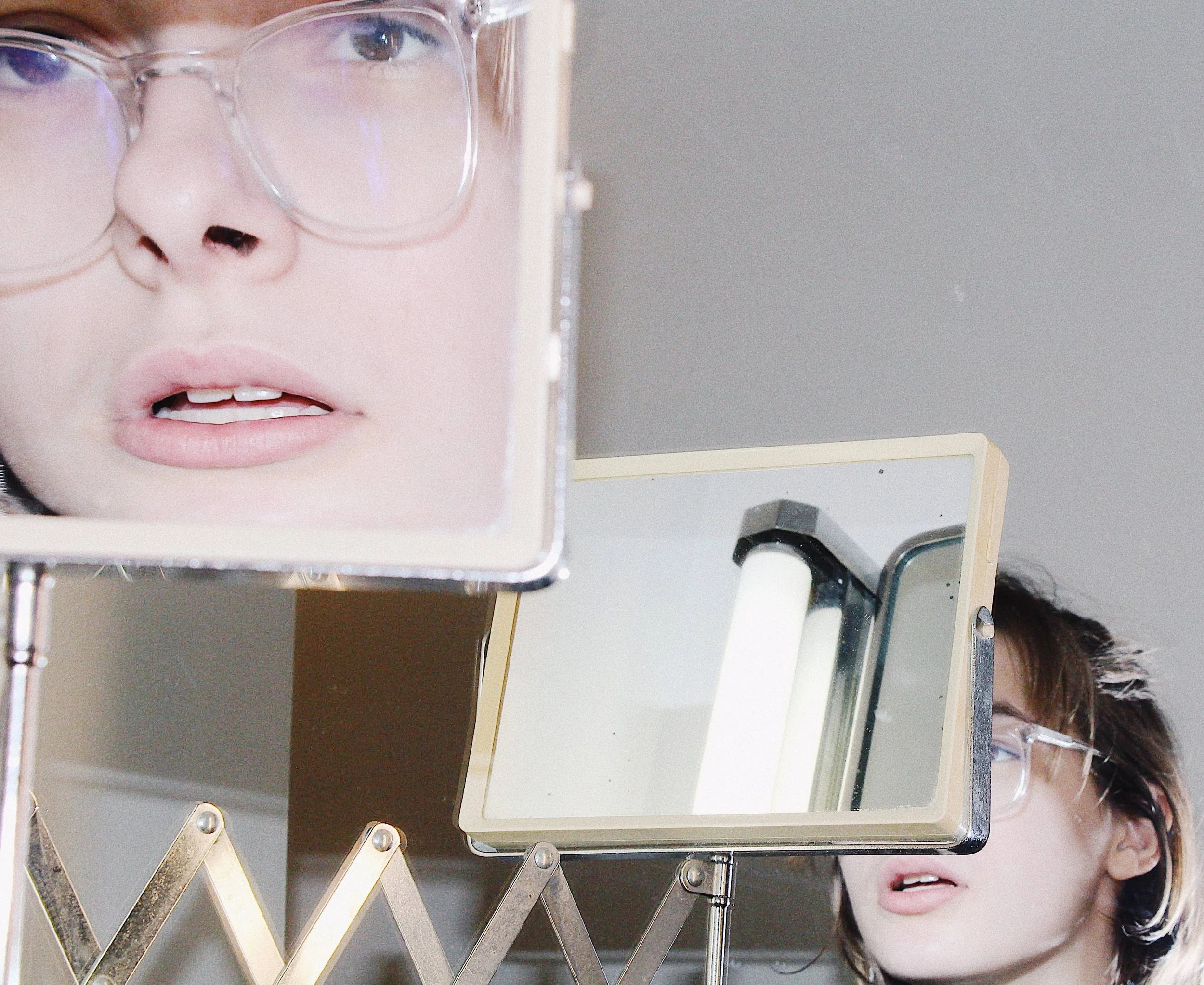 Close-up of a person's face with glasses looking into a mirror, with reflection seen in a separate smaller mirror.