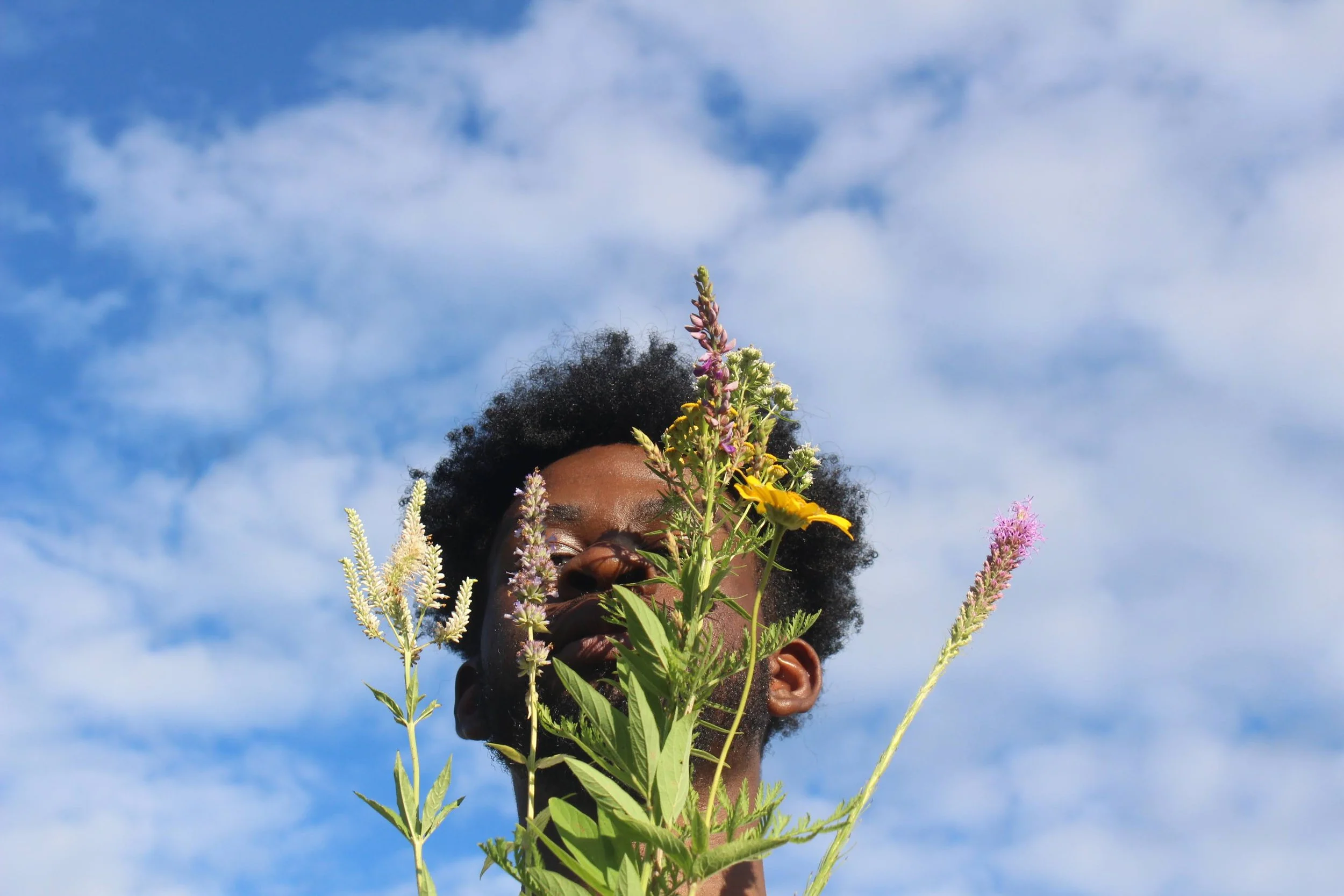 A person with curly hair holds a bouquet of colorful flowers in front of their face against a bright blue sky with scattered clouds.