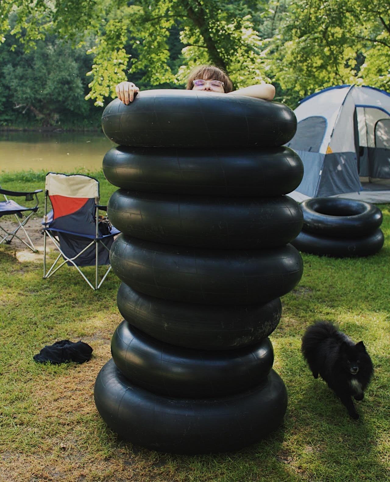 A young girl with glasses peeking over a stack of large black inner tubes outdoors near a lake, with a camping tent, chairs, and a small black dog nearby.