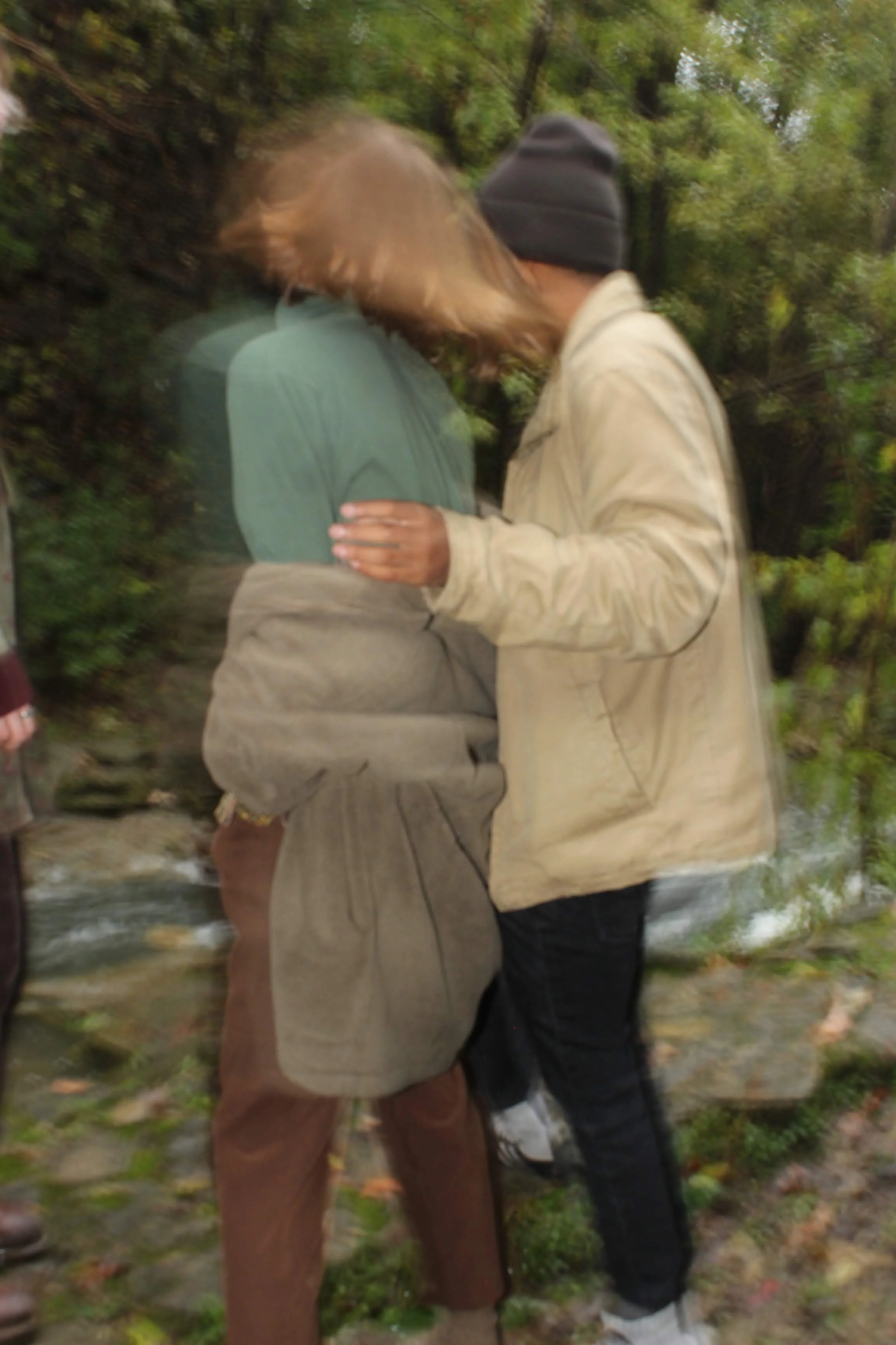 Two people standing close together outdoors, with one person touching the other's arm, near a stream with greenery in the background.