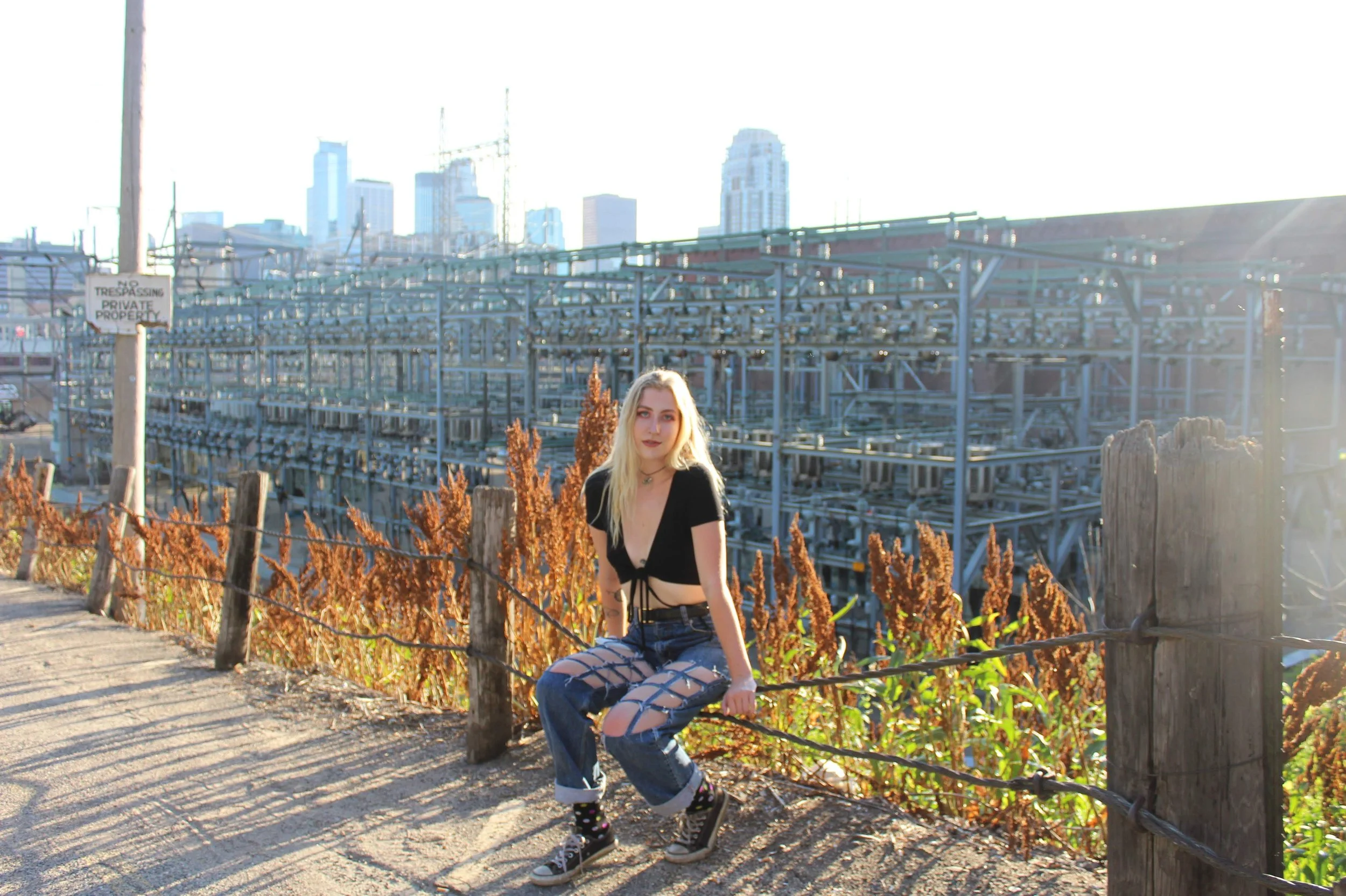A young woman with long blonde hair and a black crop top sitting on a dirt path with a city skyline in the background. She is wearing ripped jeans, platform sneakers, and starry socks. The sunlight creates a lens flare and she poses in front of a rus