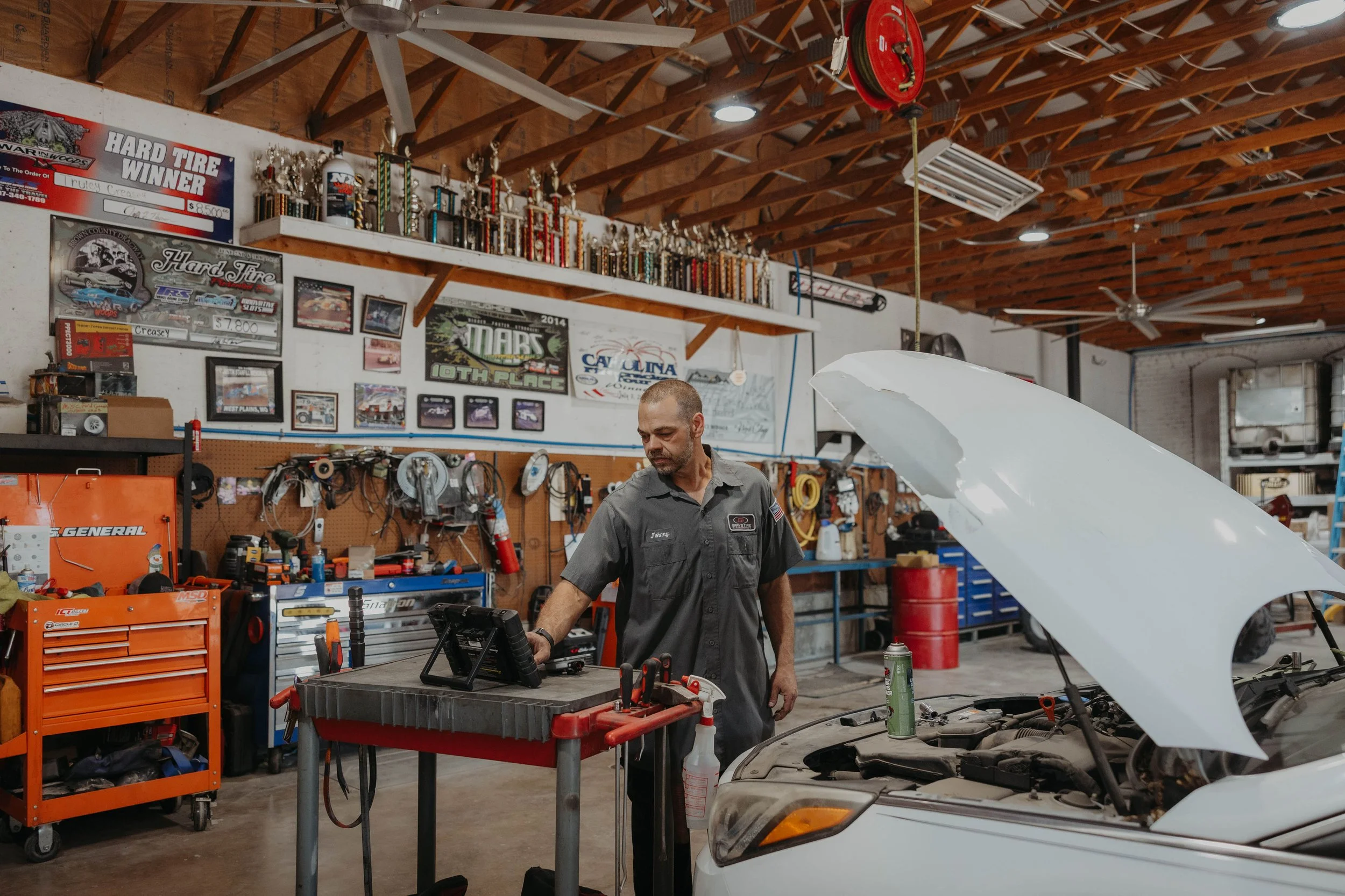 An auto mechanic in a garage working on a car's engine with the hood open. The garage has wooden ceiling beams, various trophies and posters on the walls, and tools on a pegboard and workbench.