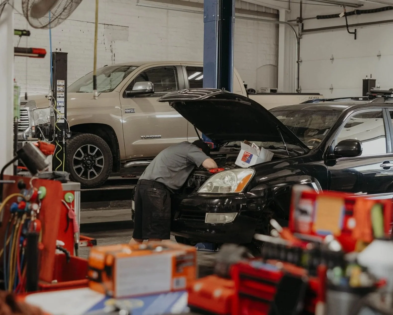 A mechanic working under the hood of a black car in an auto repair shop, with a beige pickup truck parked on a lift in the background.