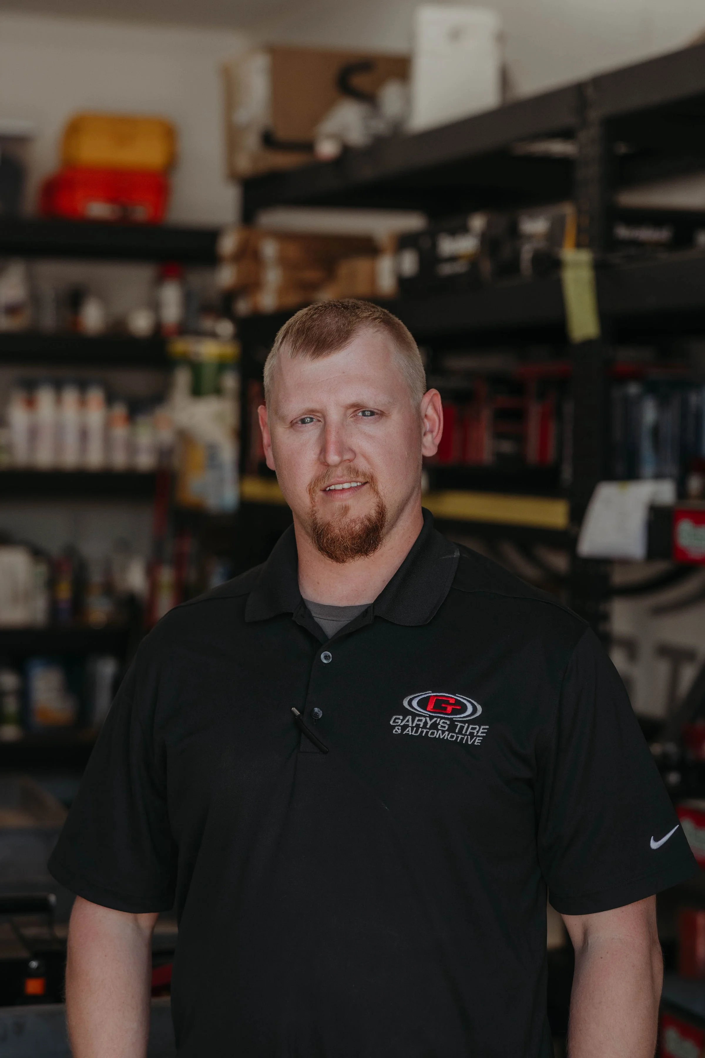 A man with short red hair and a goatee wearing a black polo shirt with the logo of Gary's Tire & Automotive.