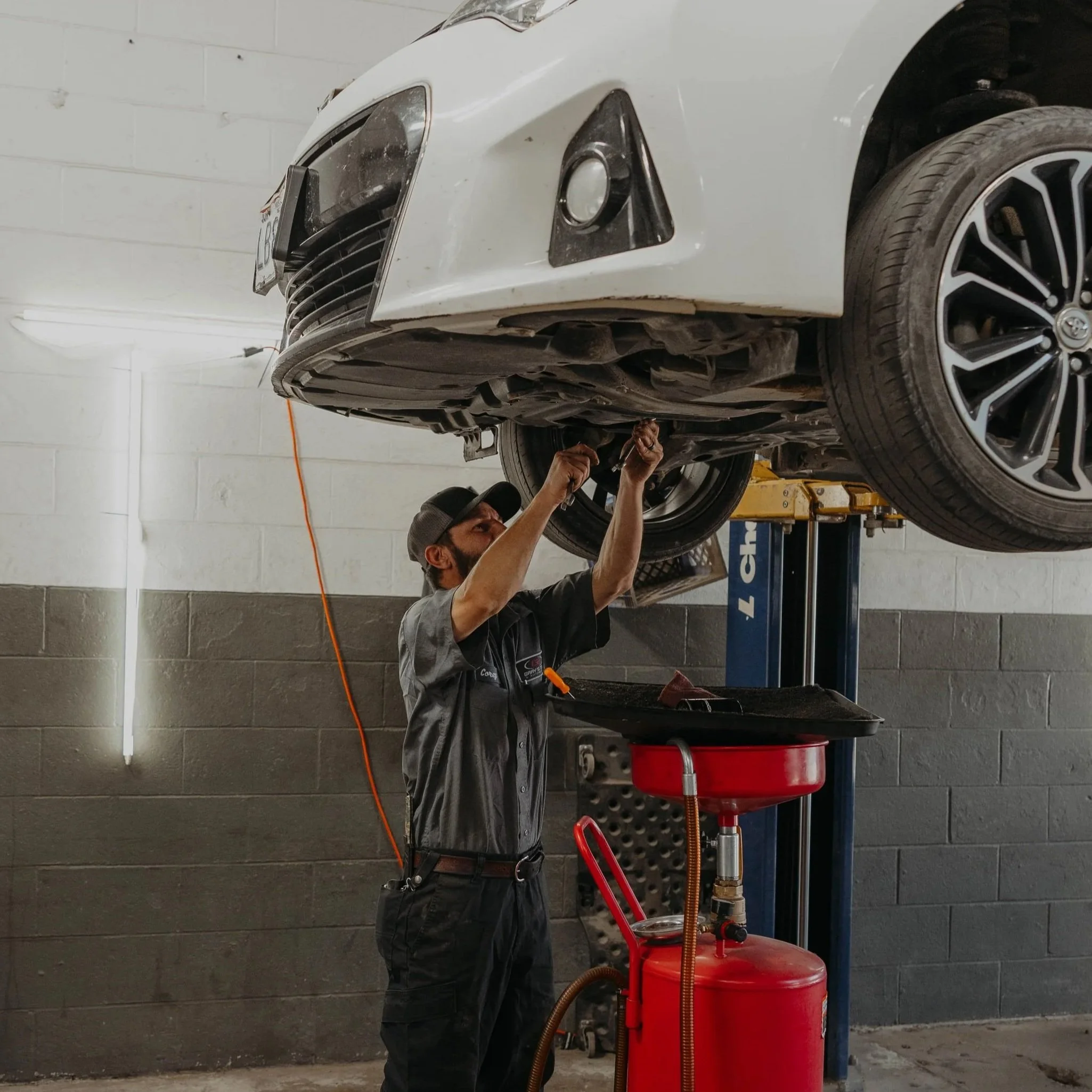 A person wearing a gray shirt and dark pants, with a hat and glasses, is standing at an automotive workshop, working under a white car that is elevated on a lift. The person appears to be using tools to repair or inspect the vehicle's underside. There is a red oil drain pan and a red storage container near the person, with an orange extension cord hanging in the background. The workshop has gray cinder block walls and fluorescent lighting.