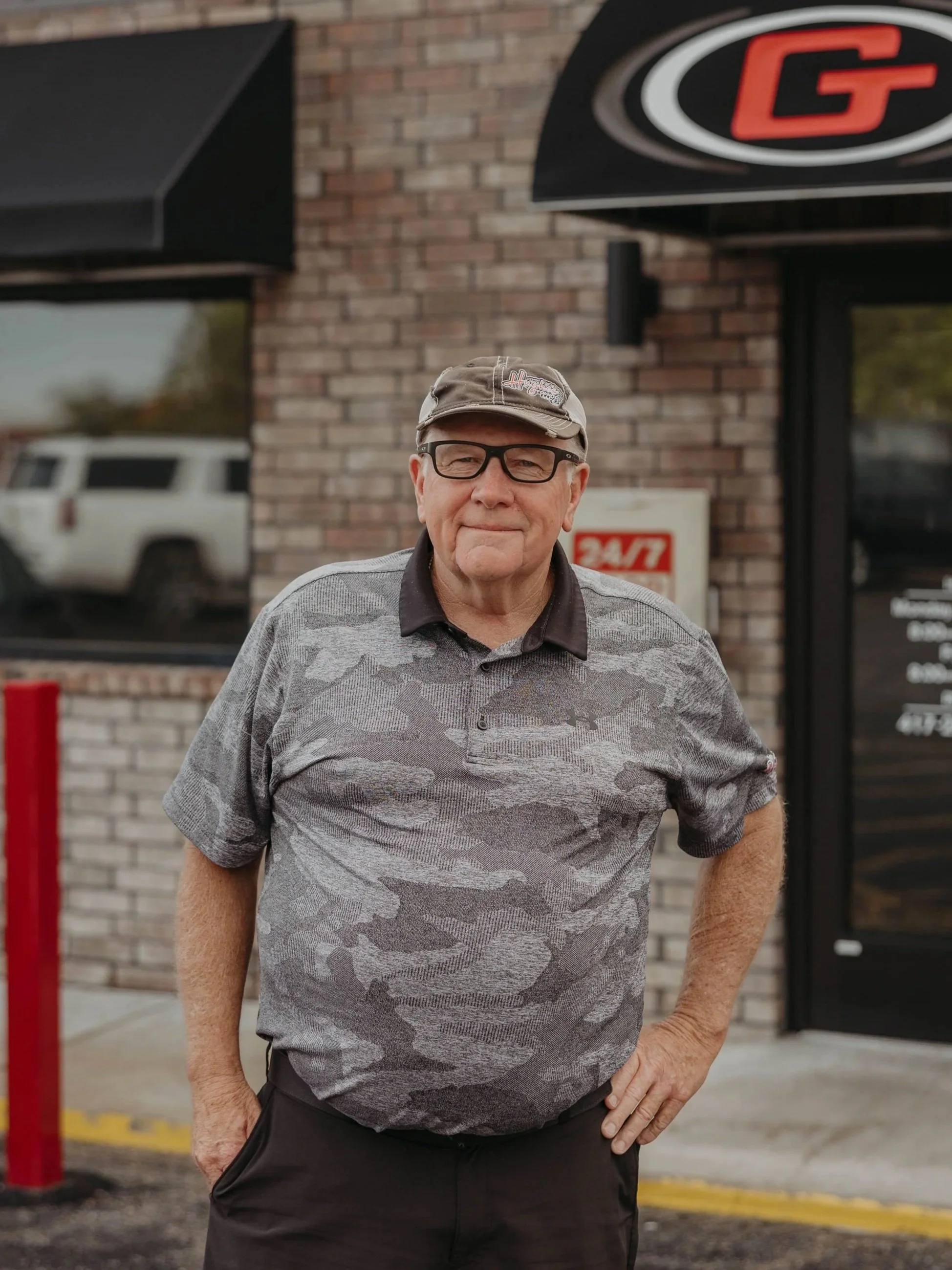 An elderly man with glasses wearing a camo cap and a gray camouflage patterned polo shirt standing outside a building with brick wall and signs, with a white vehicle reflected in the window behind him.