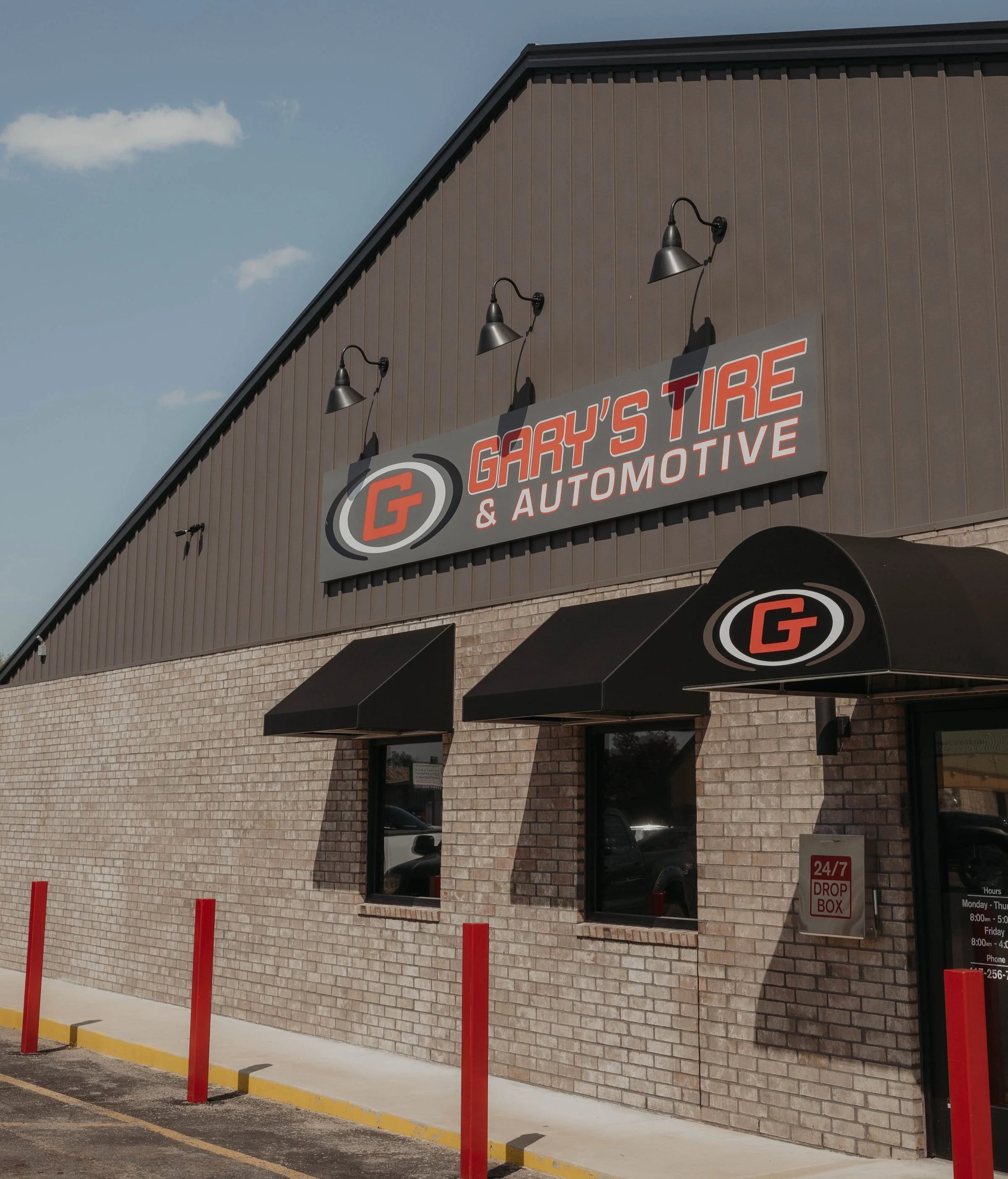 Front exterior of Gary's Tire & Automotive store with a large sign and black awnings, red parking barriers, and a brick lower wall under a blue sky with a few clouds.