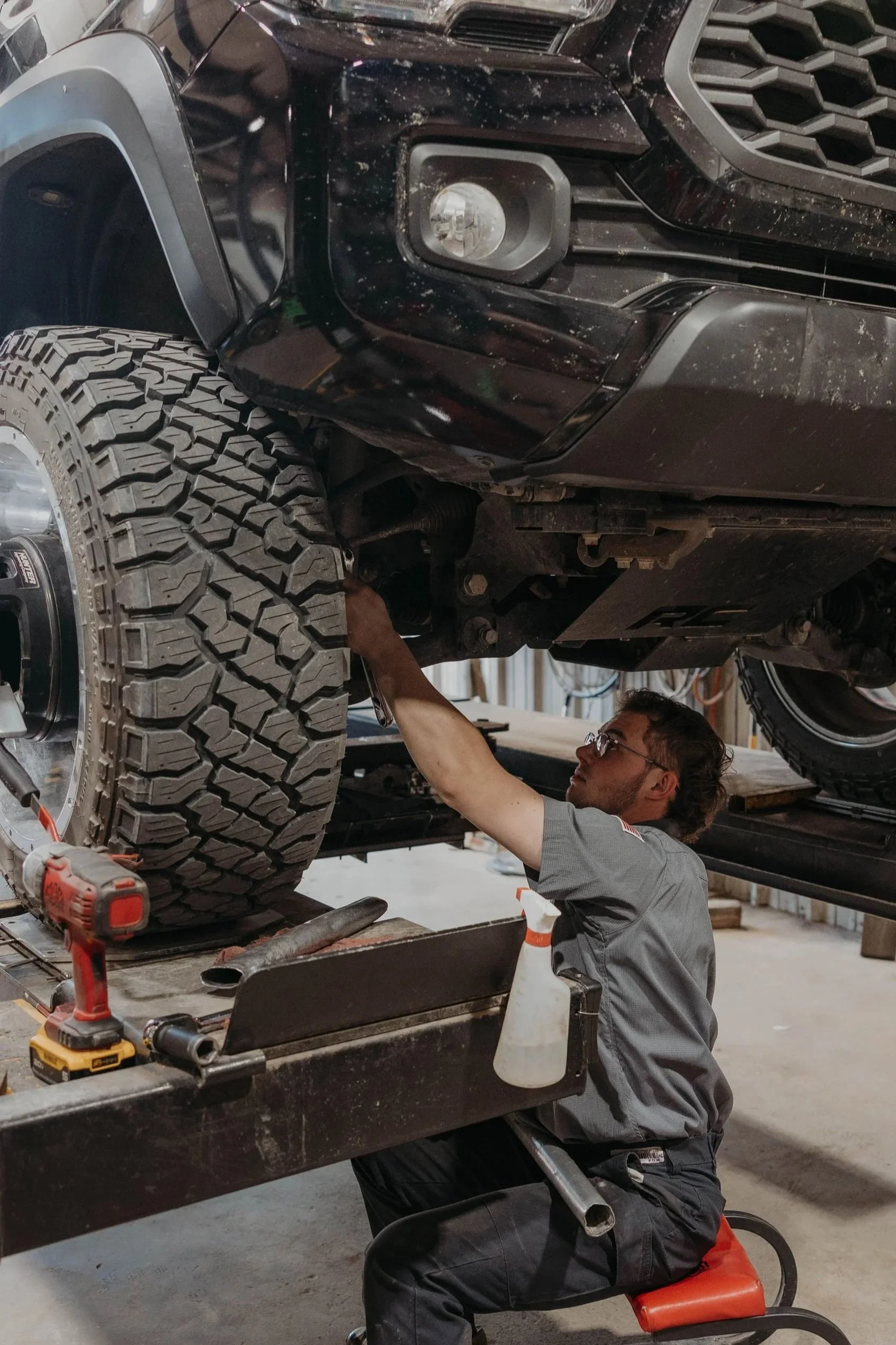 A person working underneath a lifted black vehicle, inspecting or repairing its front end. The person is seated on a small orange stool, wearing glasses and gray work clothes, with tools and equipment around.