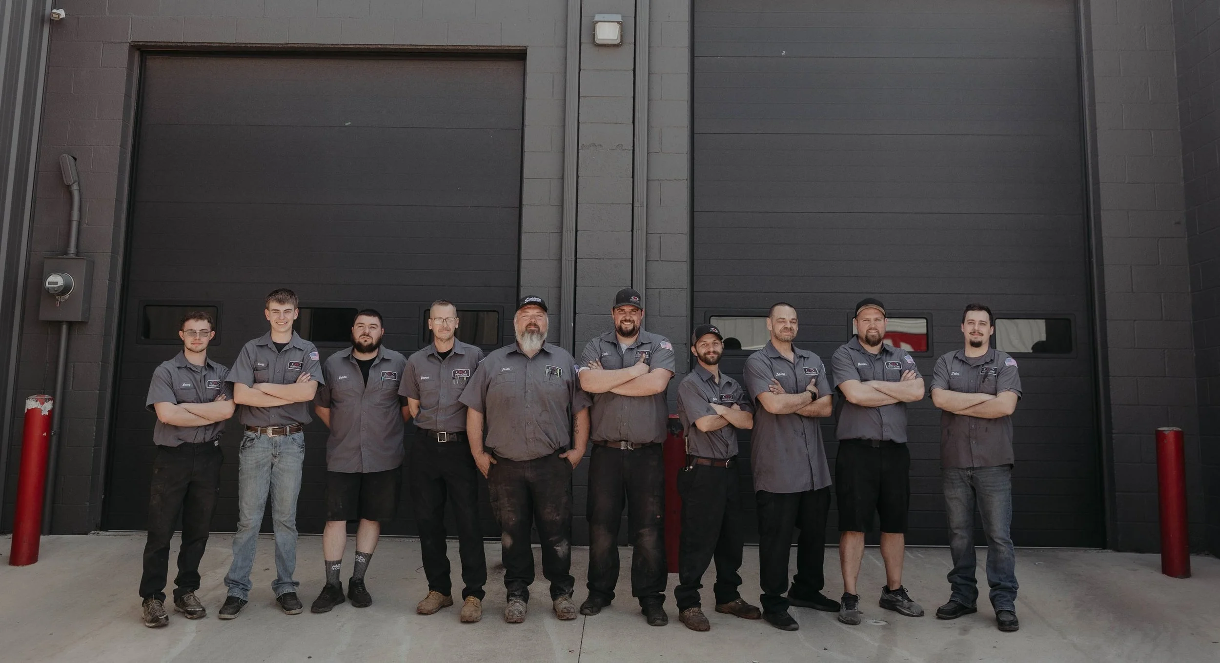 A group of ten men standing in front of a large black garage door. They are wearing gray work shirts with patches, some with company logos, and have crossed their arms or are standing with hands by their sides, smiling or looking serious.
