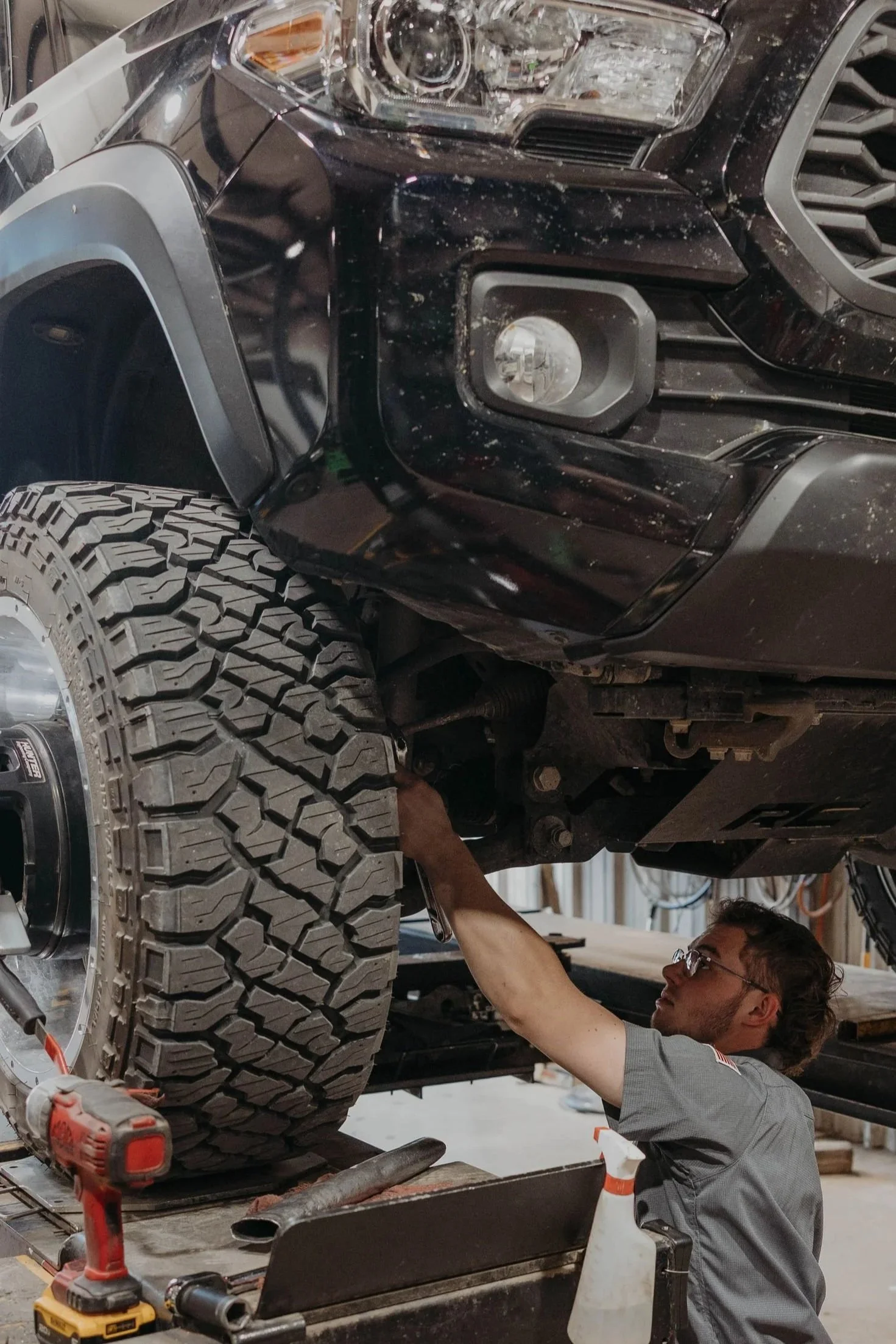 A man working underneath a lifted black vehicle, examining or repairing its suspension or undercarriage, with large off-road tires nearby in a garage or workshop.