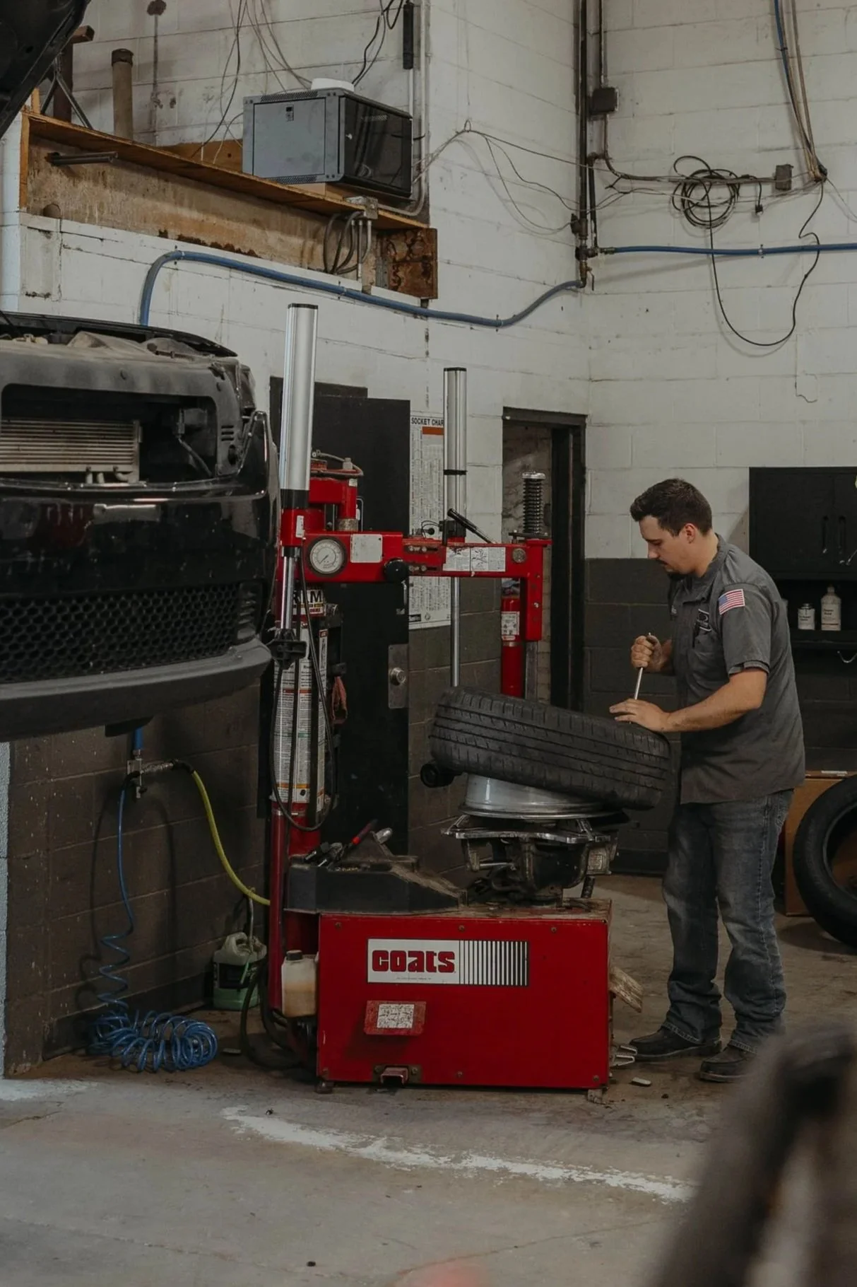 A mechanic working on a car tire mounted on a machine in an auto repair shop. The shop has exposed electrical wires and a wall-mounted TV.