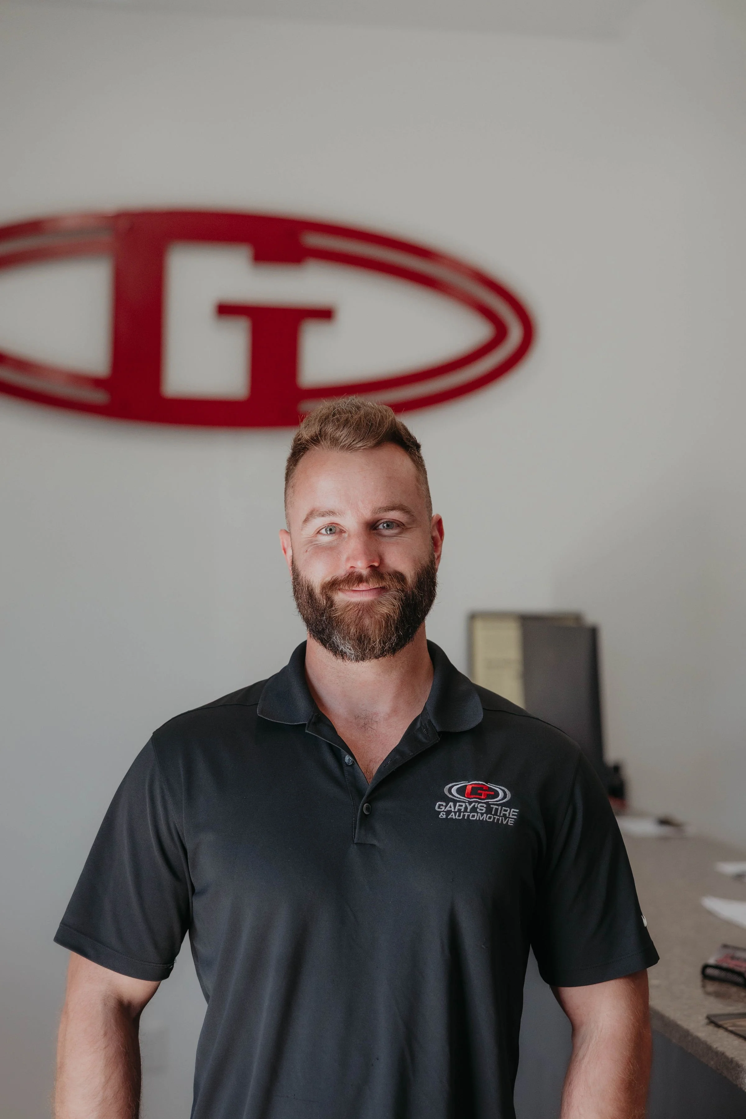 A man with a beard and short hair smiling, wearing a black polo shirt with 'Gary's Tire & Automotive' logo, standing in front of a wall with a large red and white logo.