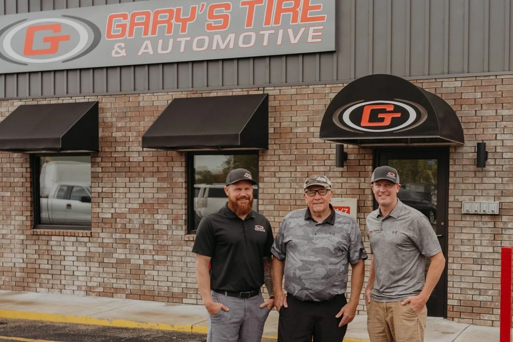 Three men standing outside Gary's Tire & Automotive shop in front of the brick building with signage and black awnings. They are smiling and dressed in casual and branded work attire.