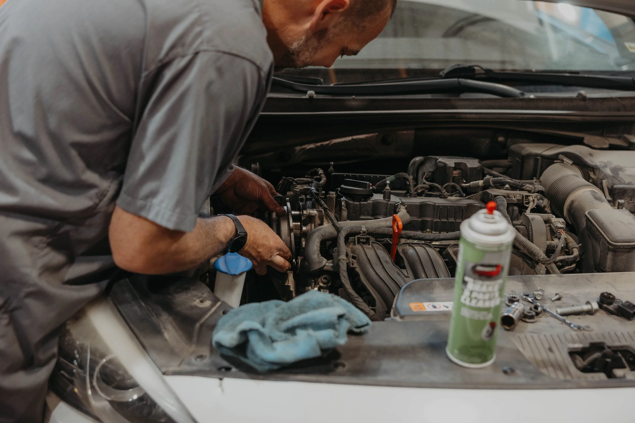 A mechanic working on a car engine in a garage, with various tools and supplies on the workbench.