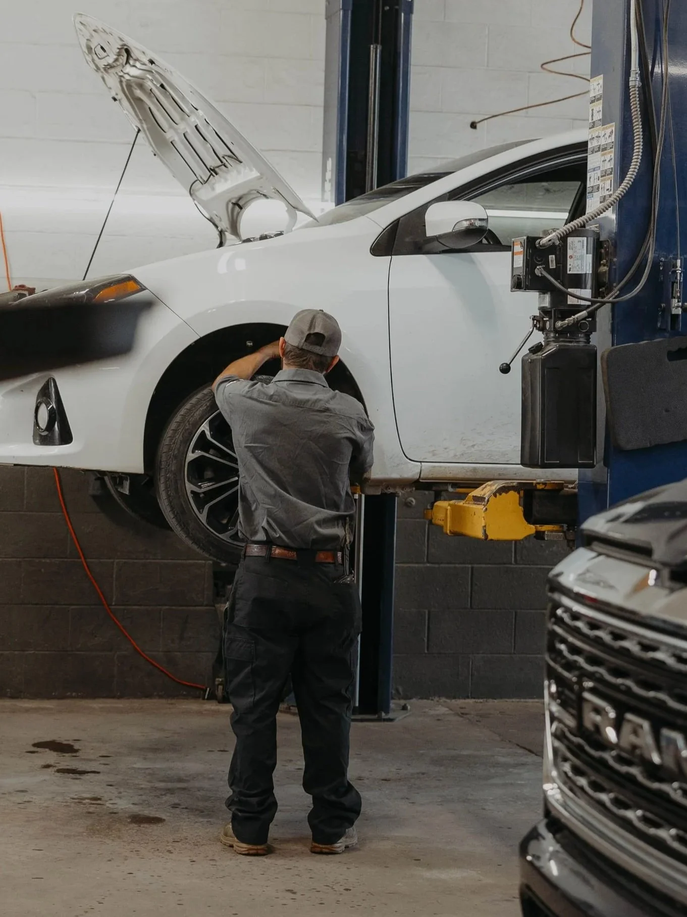 An auto mechanic working on the front left wheel of a white car lifted on a hydraulic service lift. The mechanic is wearing a gray uniform and a gray cap, with tools in hand. The shop has a concrete floor with some oil stains and a black cinder block wall in the background.