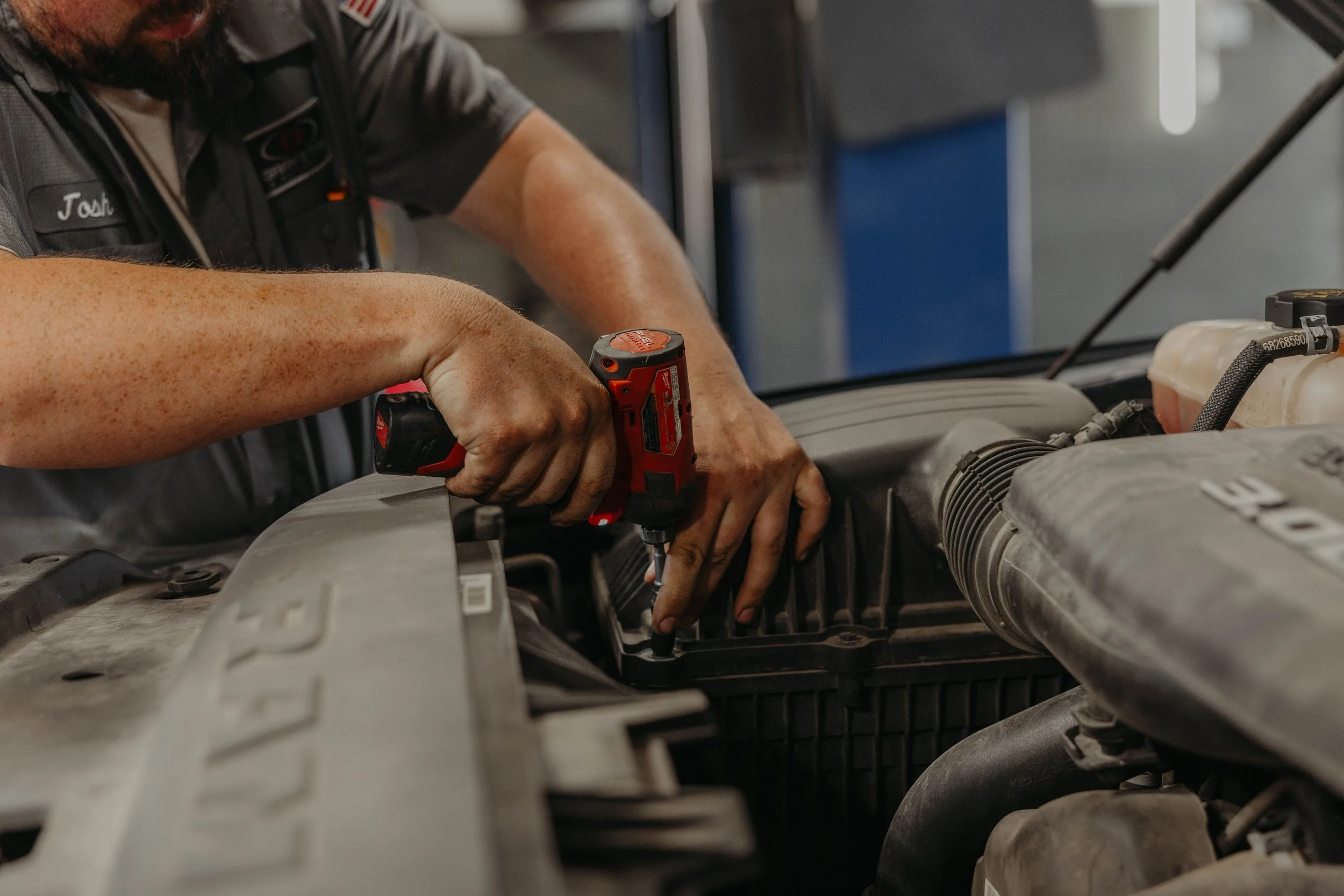 A person wearing a dark work uniform is using a cordless drill to work on a vehicle engine. The person is focused on the task, and the engine bay contains various mechanical components and tools.