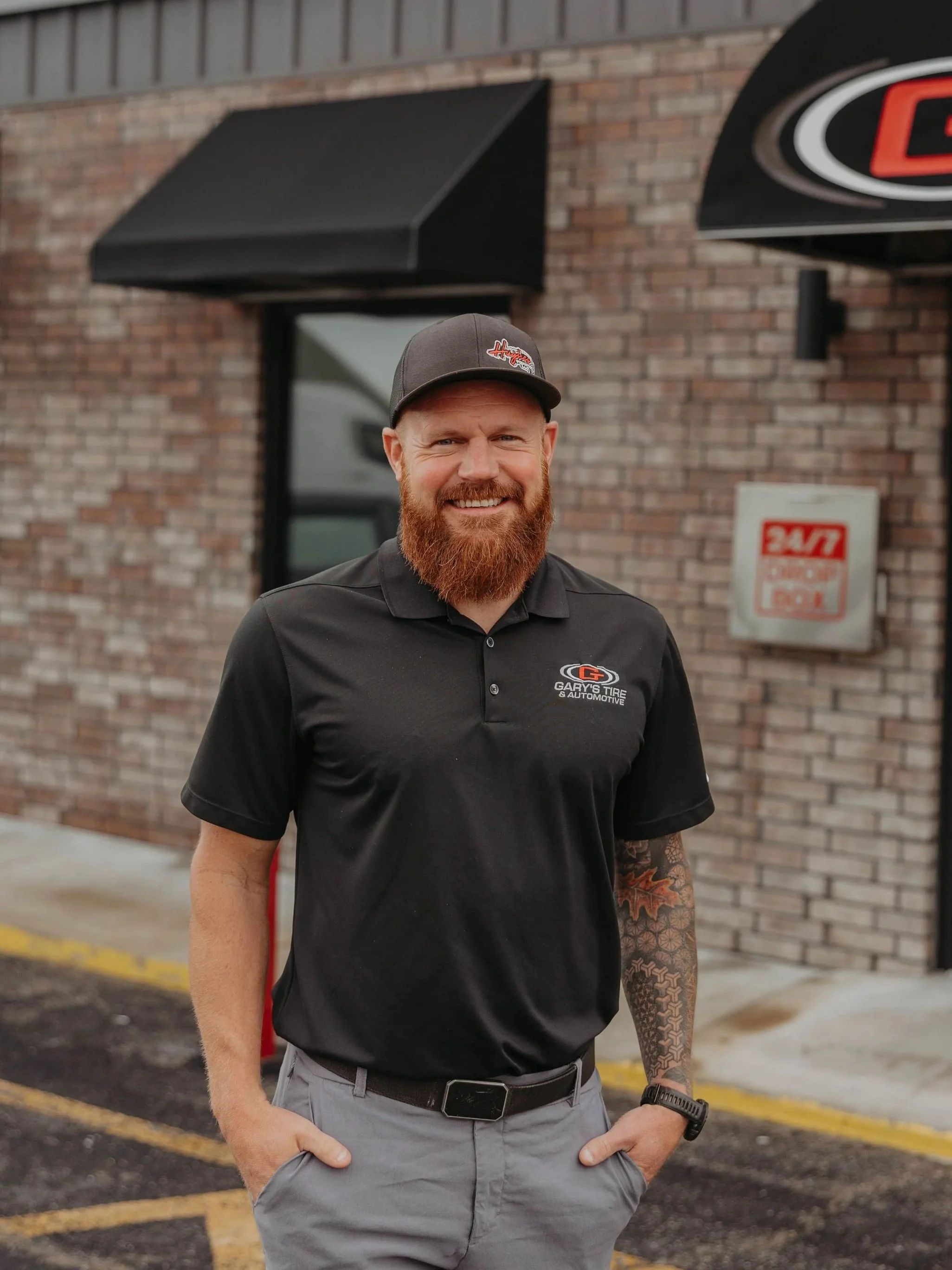 Smiling man with red beard and tattooed arm, wearing a black Gary's Tire & Automotive polo shirt, cap, and watch, standing outdoors in front of a brick building with black awnings and parking lot.