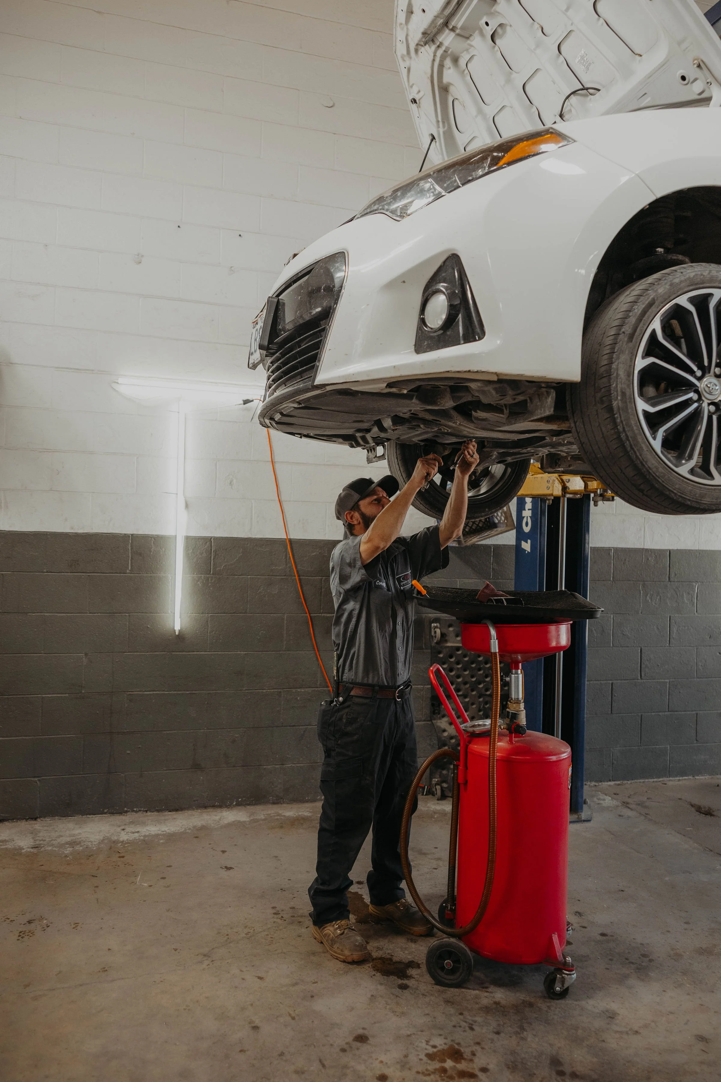 A mechanic working underneath a white car that is lifted on a hydraulic lift in an auto repair shop. The mechanic is using tools and there is a red oil drain container nearby.