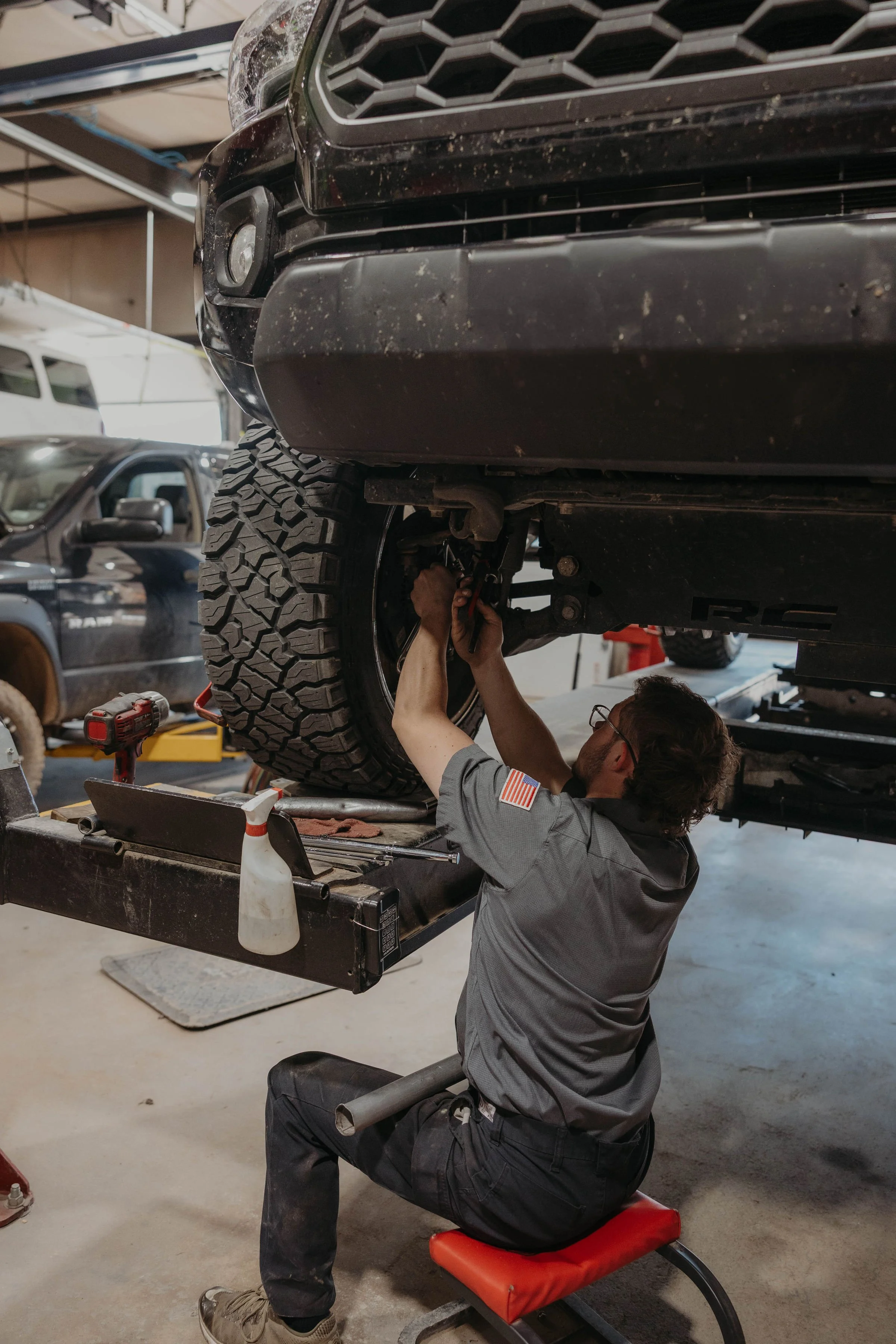 A mechanic working underneath a raised vehicle in an auto repair shop. The mechanic is using tools to work on the vehicle's suspension or axle area, and the vehicle's front tire is visible. The shop has other vehicles and equipment in the background.