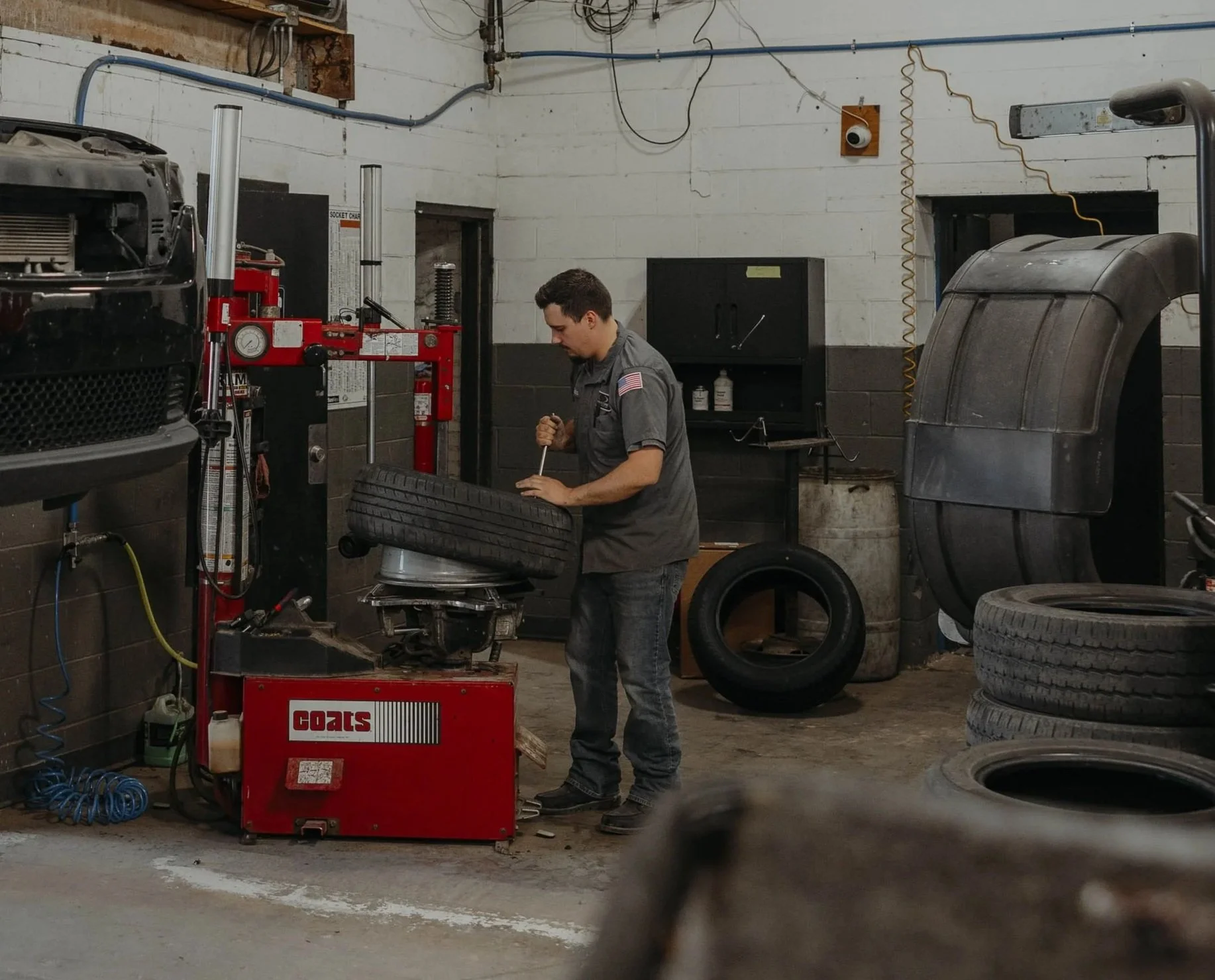 A mechanic working on a car tire in an auto repair shop, surrounded by tires, tools, and equipment.