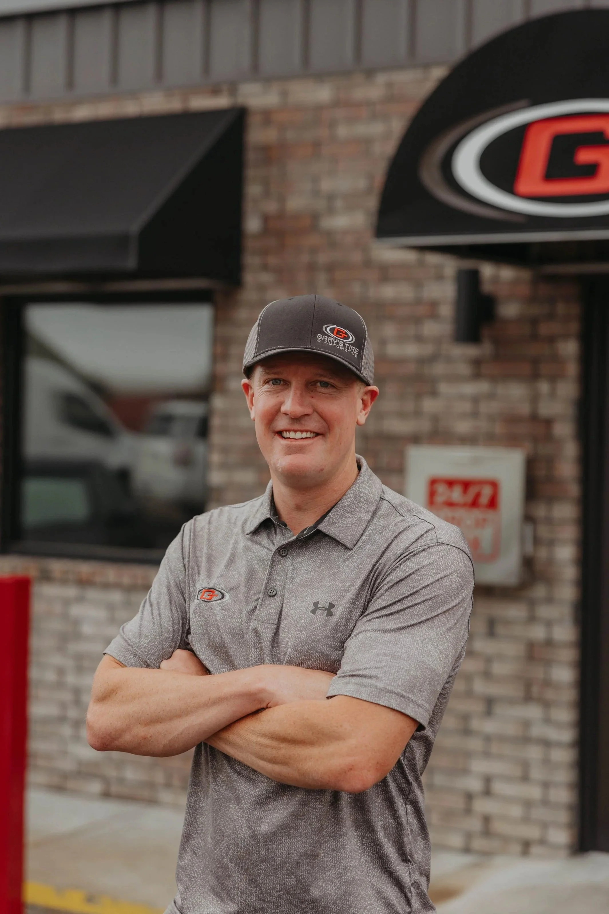 A man standing outdoors with arms crossed, smiling, wearing a gray cap and gray Under Armour shirt, in front of a brick building with a Gary Tire logo sign.