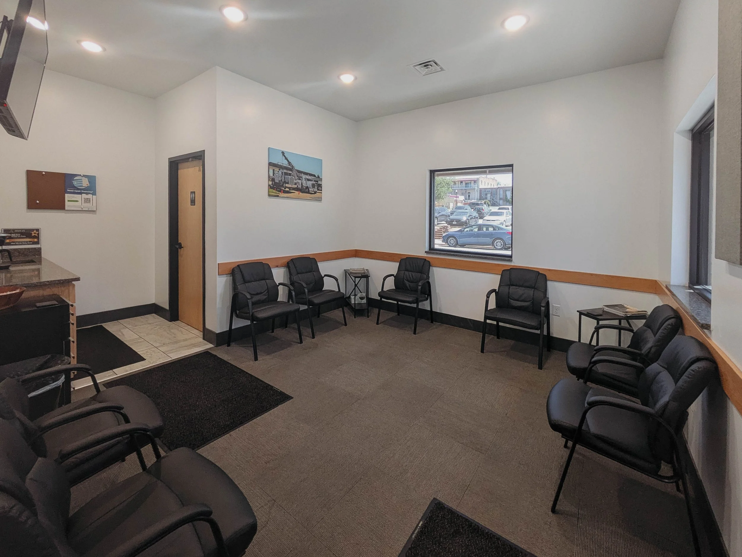 A waiting room with black chairs arranged along the walls, a window showing a parking lot outside, and a few posters on the walls.